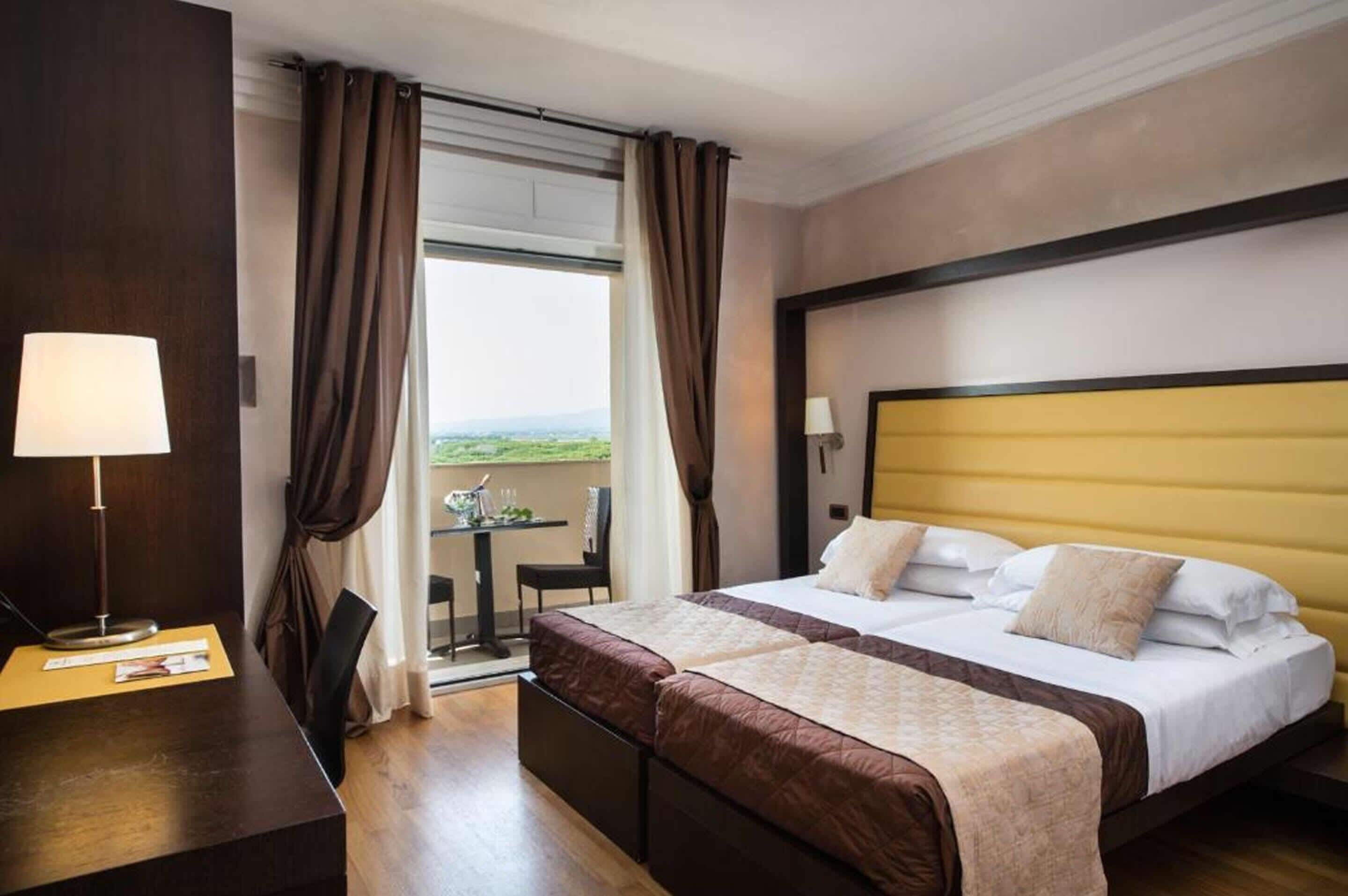 A hotel room with two beds, a desk, a chair, and a view of the outdoors.