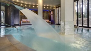 A modern indoor pool with a water slide, a lounge area with a sofa, and large windows offering a view of the outdoors. - Park Hotel Marinetta (Bibbona)