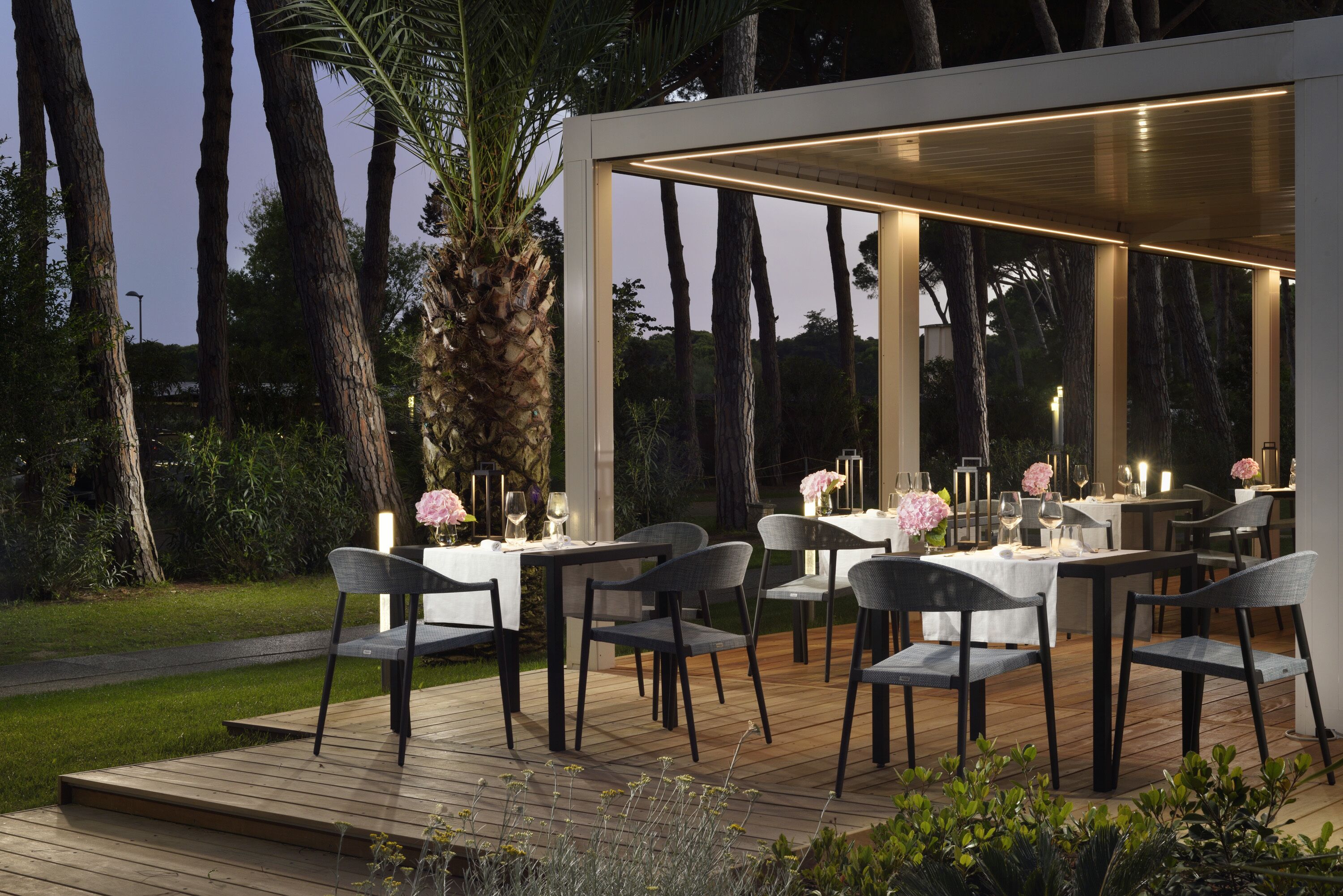 An outdoor dining area with tables and chairs, surrounded by trees and a wooden deck.