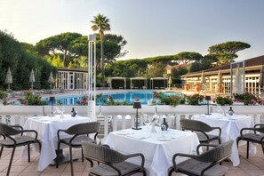 Un área de comedor al aire libre con mesas y sillas, una piscina y abundante vegetación de fondo.