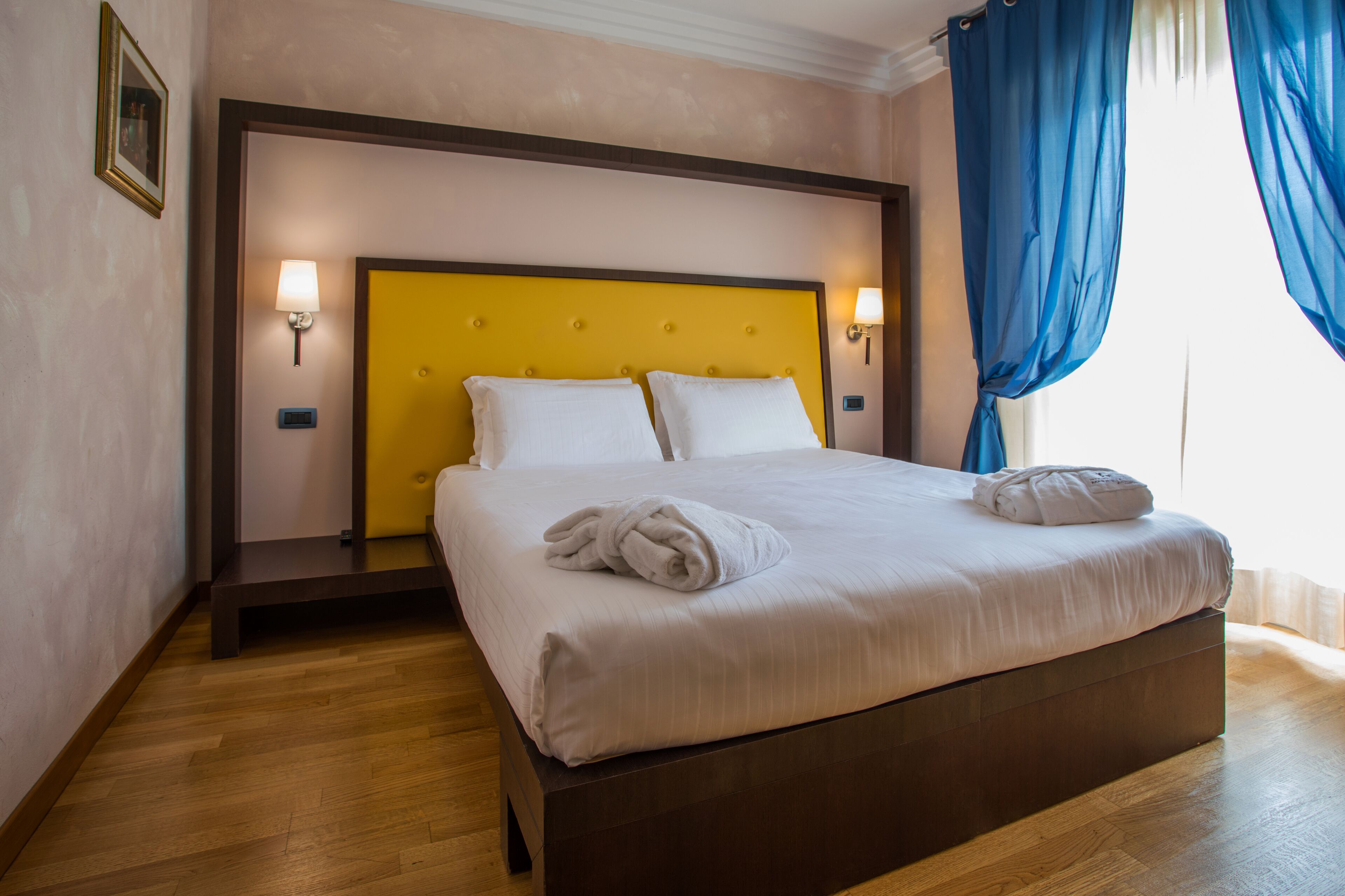 A hotel room with a wooden floor, a yellow headboard, white bedding, and a blue curtain.