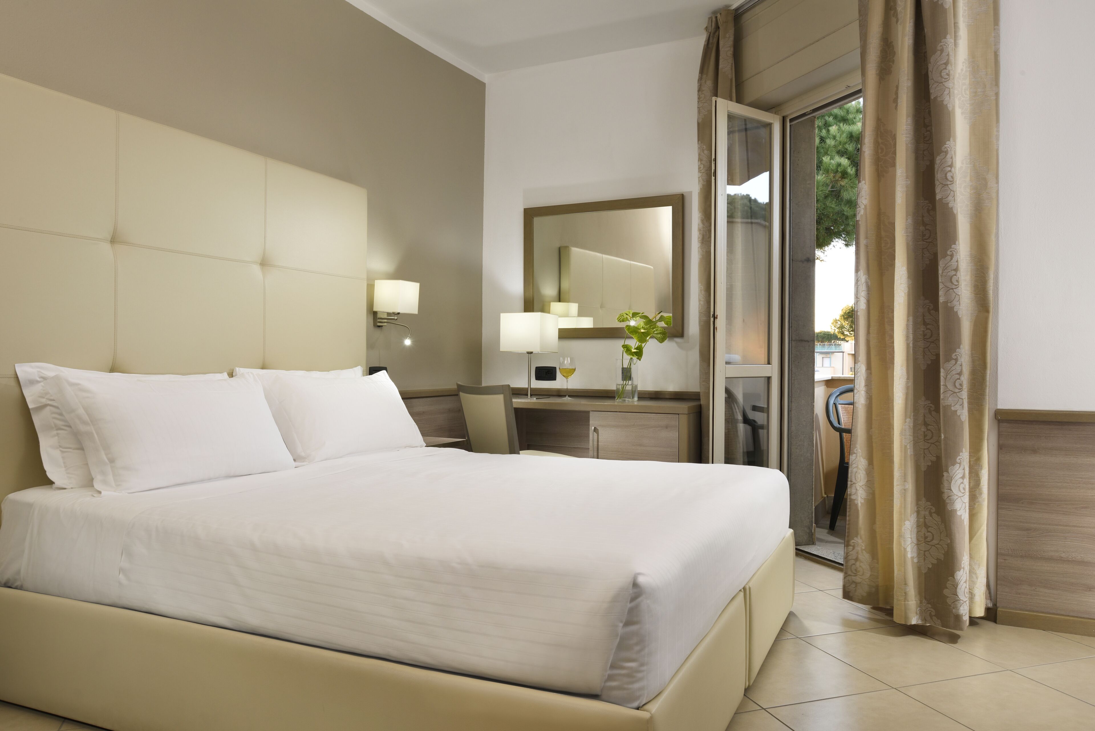 A hotel room with a large bed, a desk, a mirror, and a view of the outdoors.