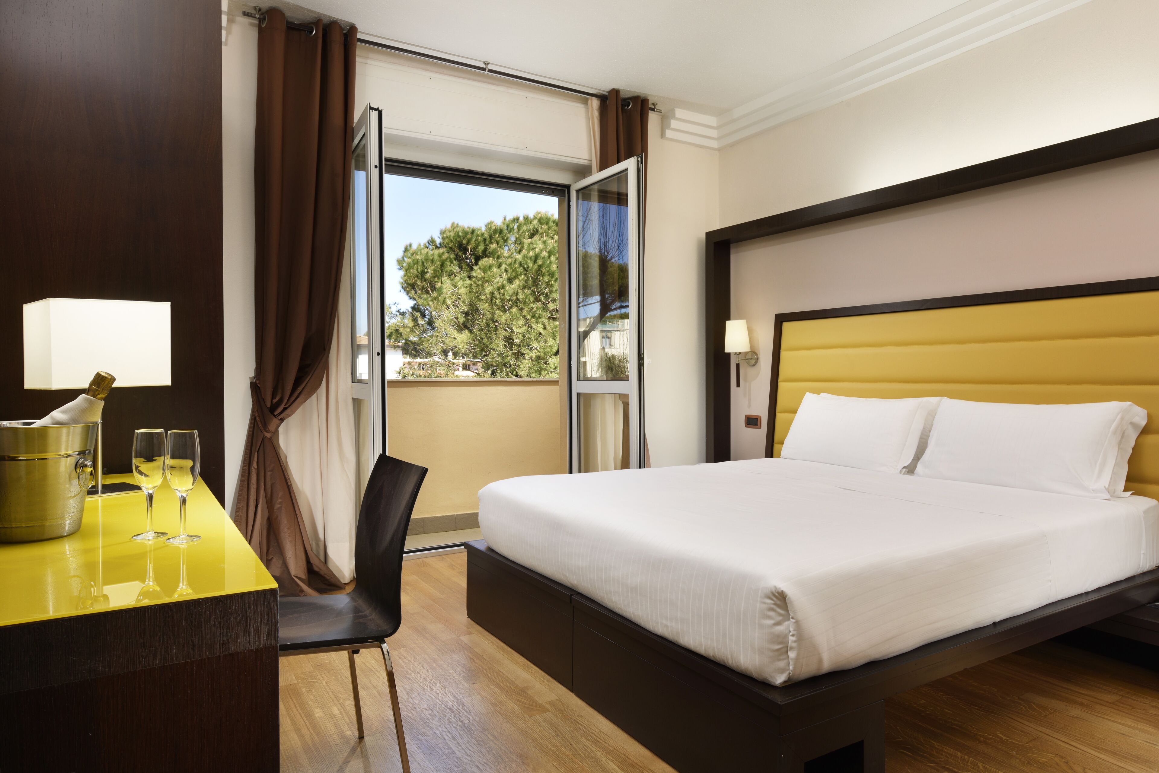 Una habitación de hotel con cama, escritorio con lámpara, silla y ventana con cortinas.