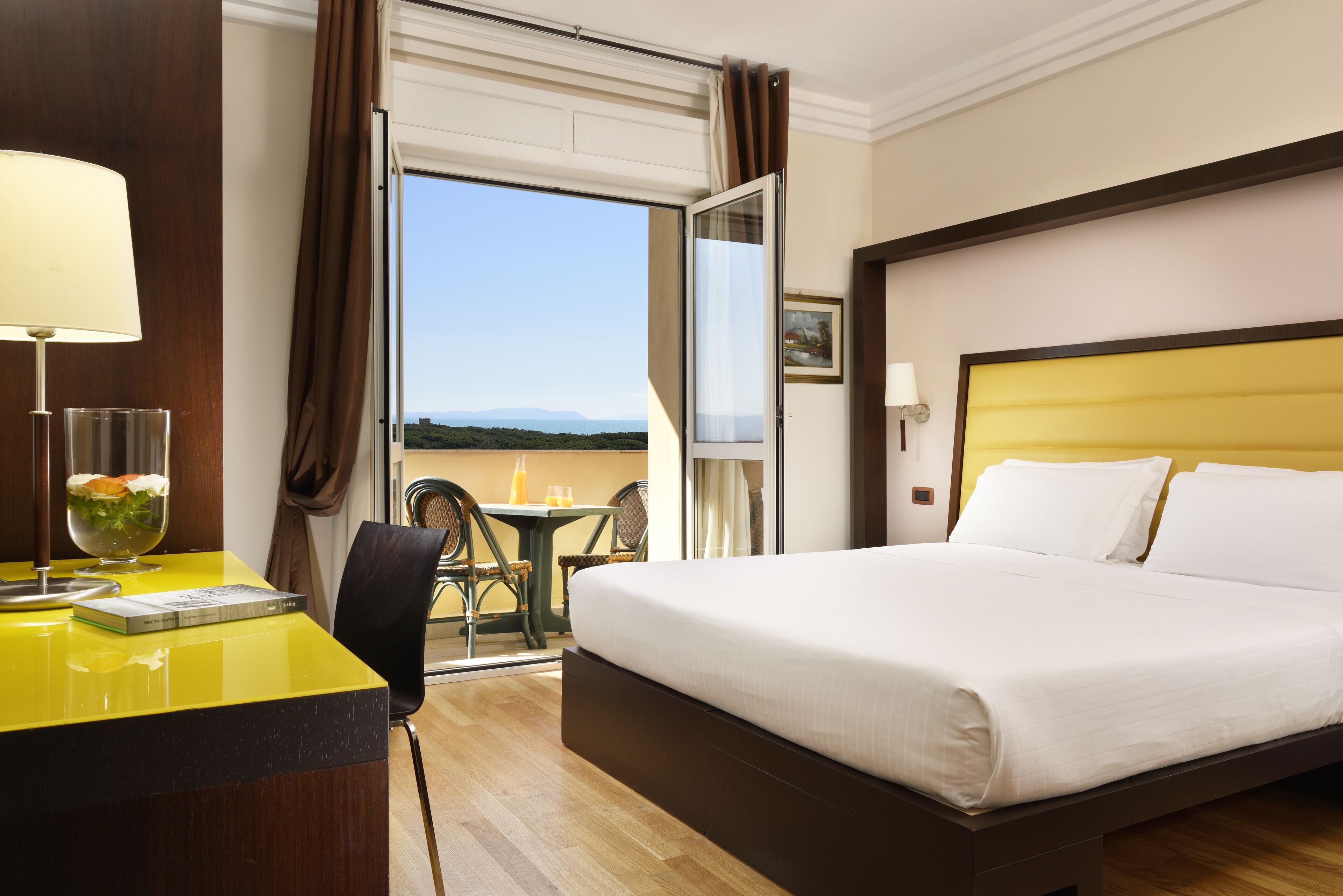 Habitación de hotel con cama, escritorio con lámpara, silla, mesa con una bebida y vistas al exterior.