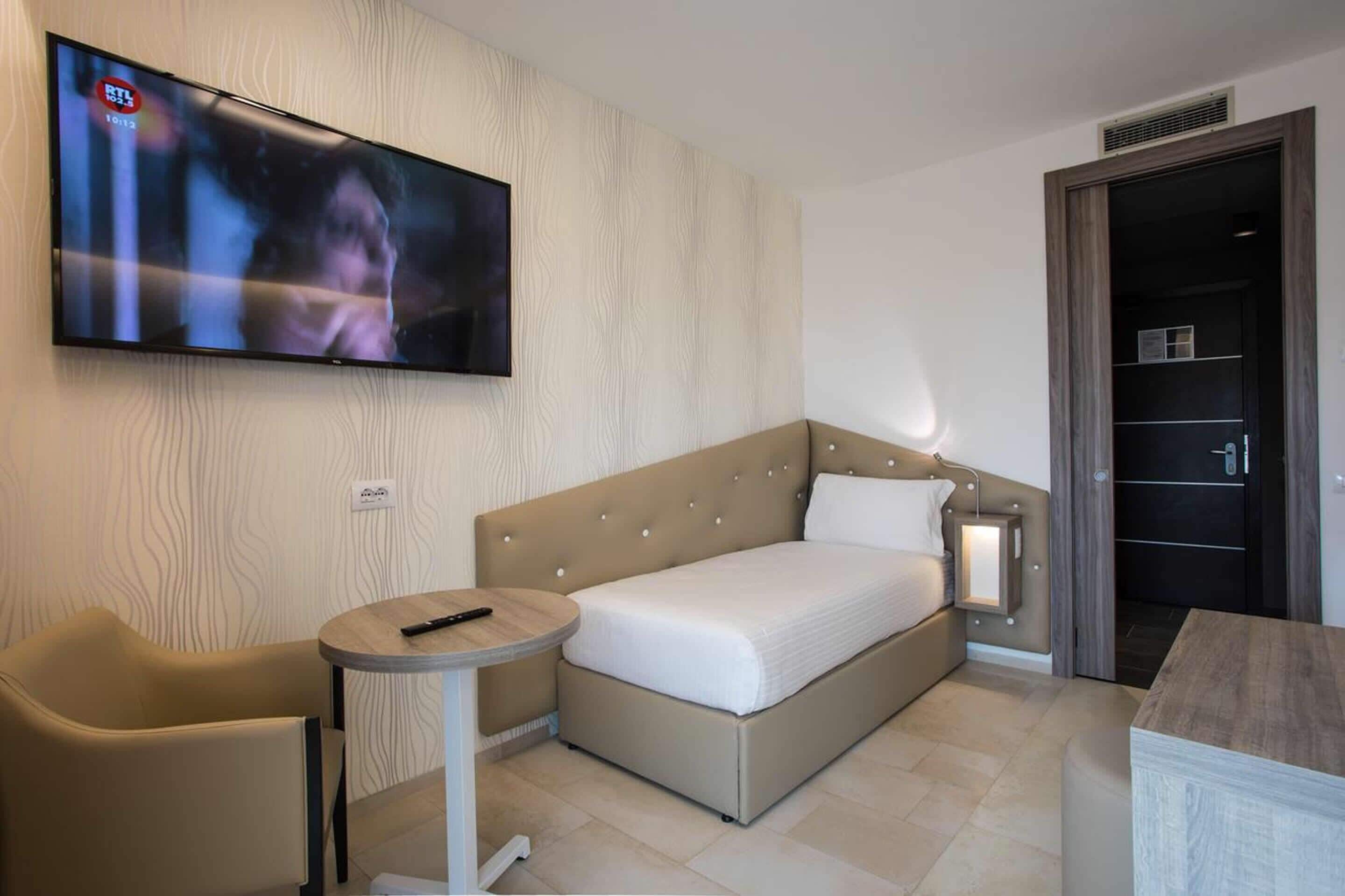 Moderni hotellihuone, jossa on sänky, seinälle asennettu televisio, pieni pyöreä pöytä ja tuoli.