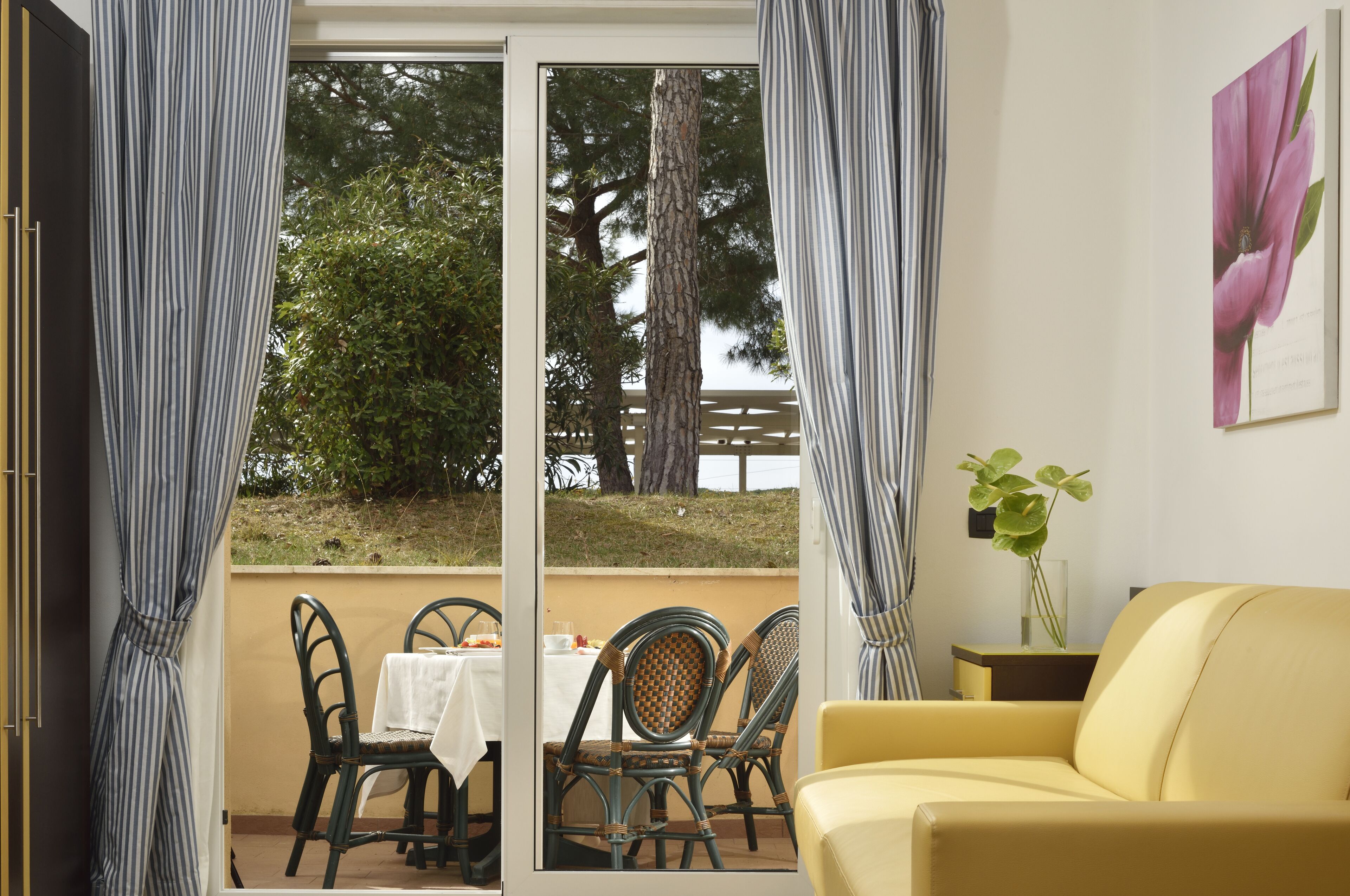 Quarto com um sofá amarelo, uma mesa de jantar com cadeiras e vista para árvores e uma cerca através de uma porta de vidro.