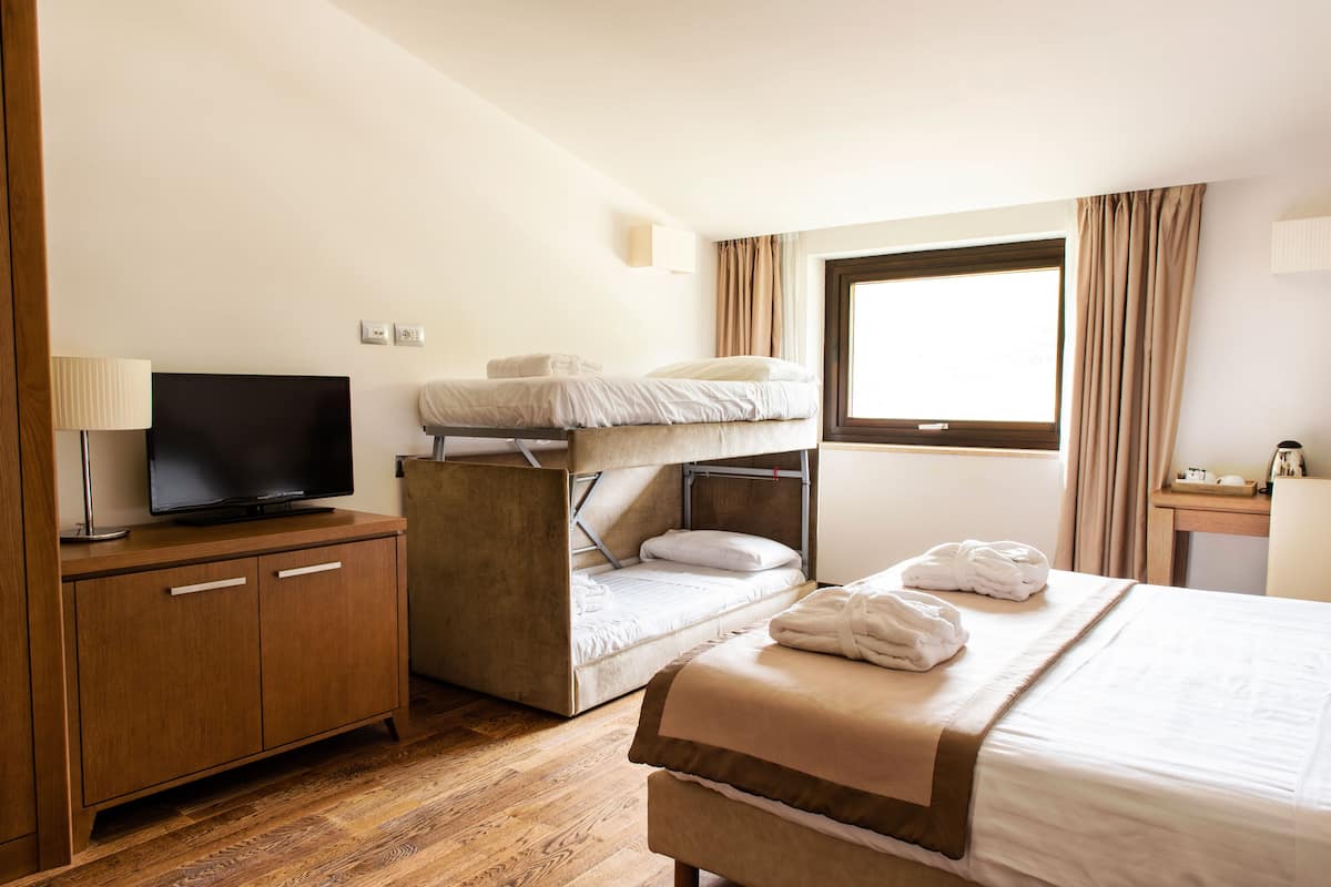 Ein Hotelzimmer mit einem Etagenbett, einem Fernseher auf einer Holzkommode, einem Nachttisch mit Lampe und einem großen Fenster mit Vorhängen.