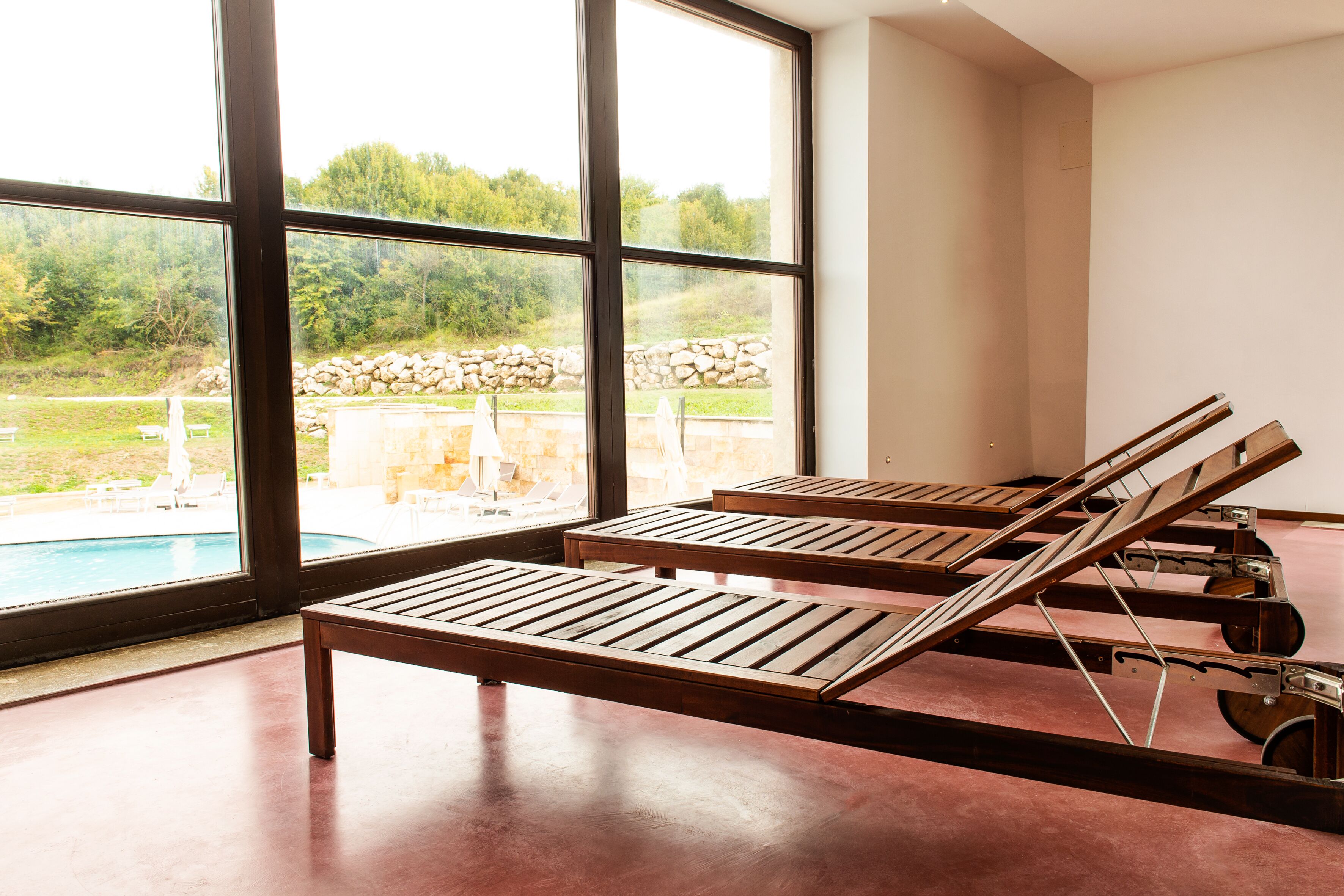 A room with large windows overlooking a swimming pool, wooden loungers, and a stone wall.
