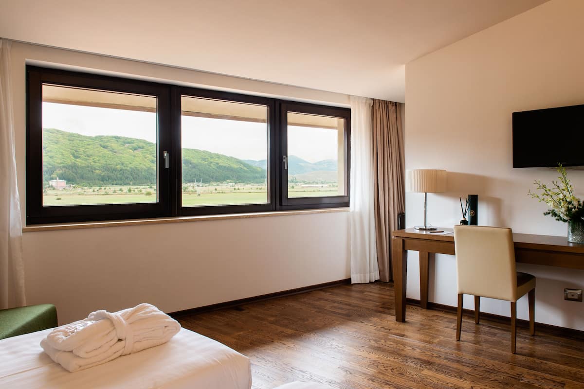 Ein Hotelzimmer mit großem Fenster, Schreibtisch, Stuhl, Fernseher und einem Bett mit zusammengelegtem Handtuch.