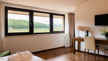 A hotel room with a large window, a desk, a chair, a TV, and a bed with a rolled-up towel.