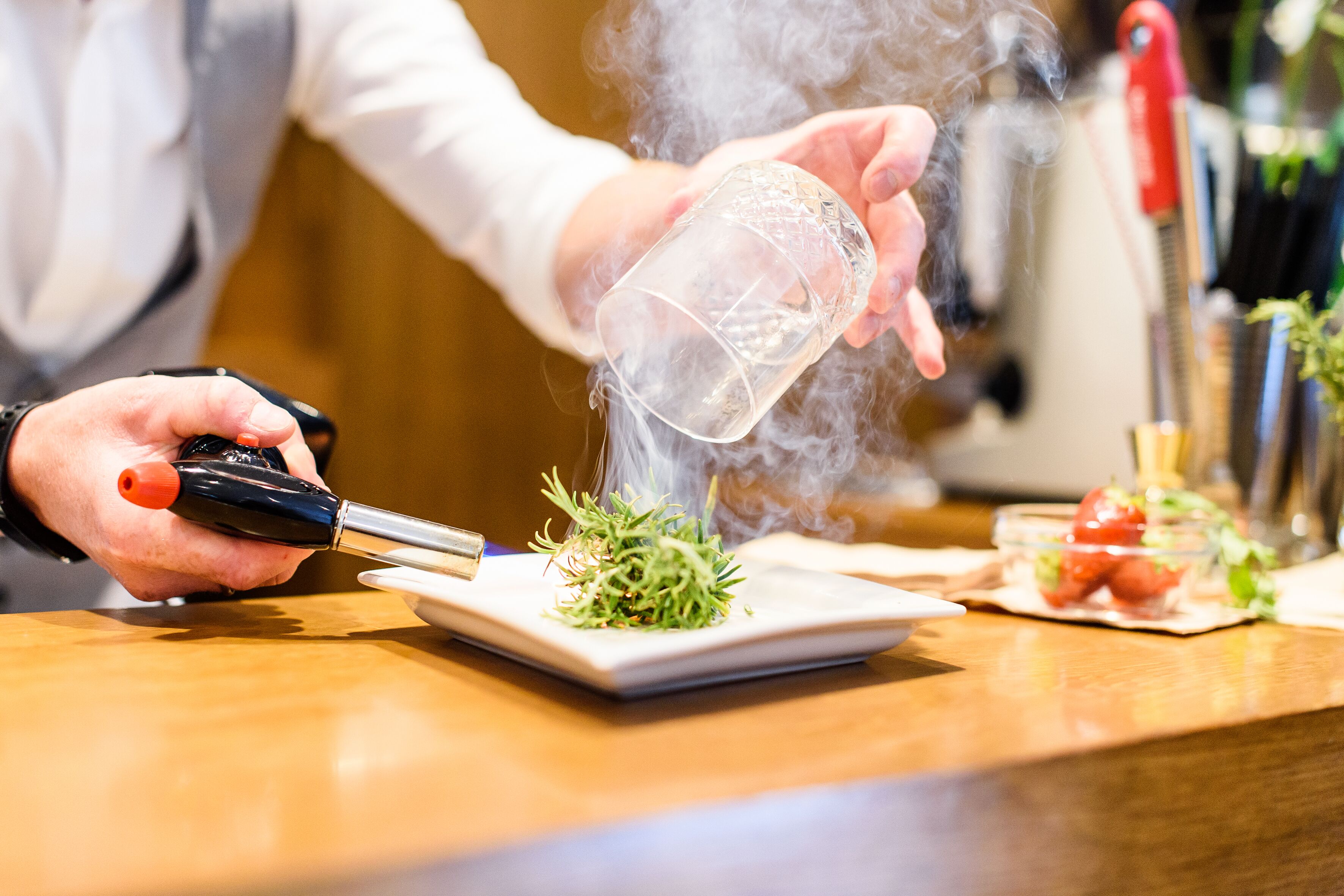 Una persona preparando un plato con un utensilio humeante, un plato y un cuenco pequeño de ingredientes sobre una mesa de madera.