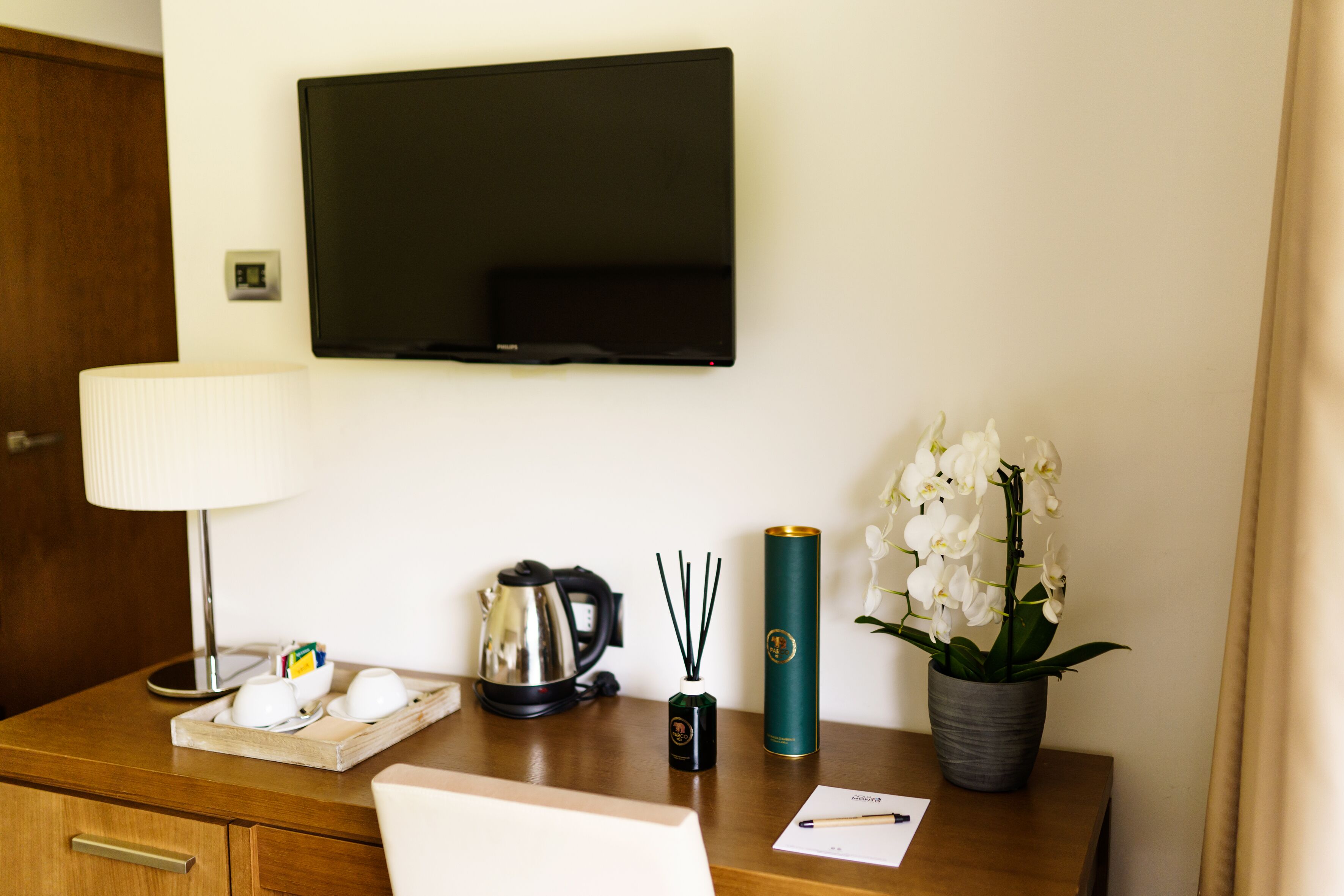 Habitación de hotel con un escritorio de madera, un televisor de pantalla plana en la pared, una lámpara, un hervidor eléctrico, una bandeja con tazas y una caja de pañuelos, una botella de difusor de caña y un ambientador en aerosol, además de una orquídea en maceta.