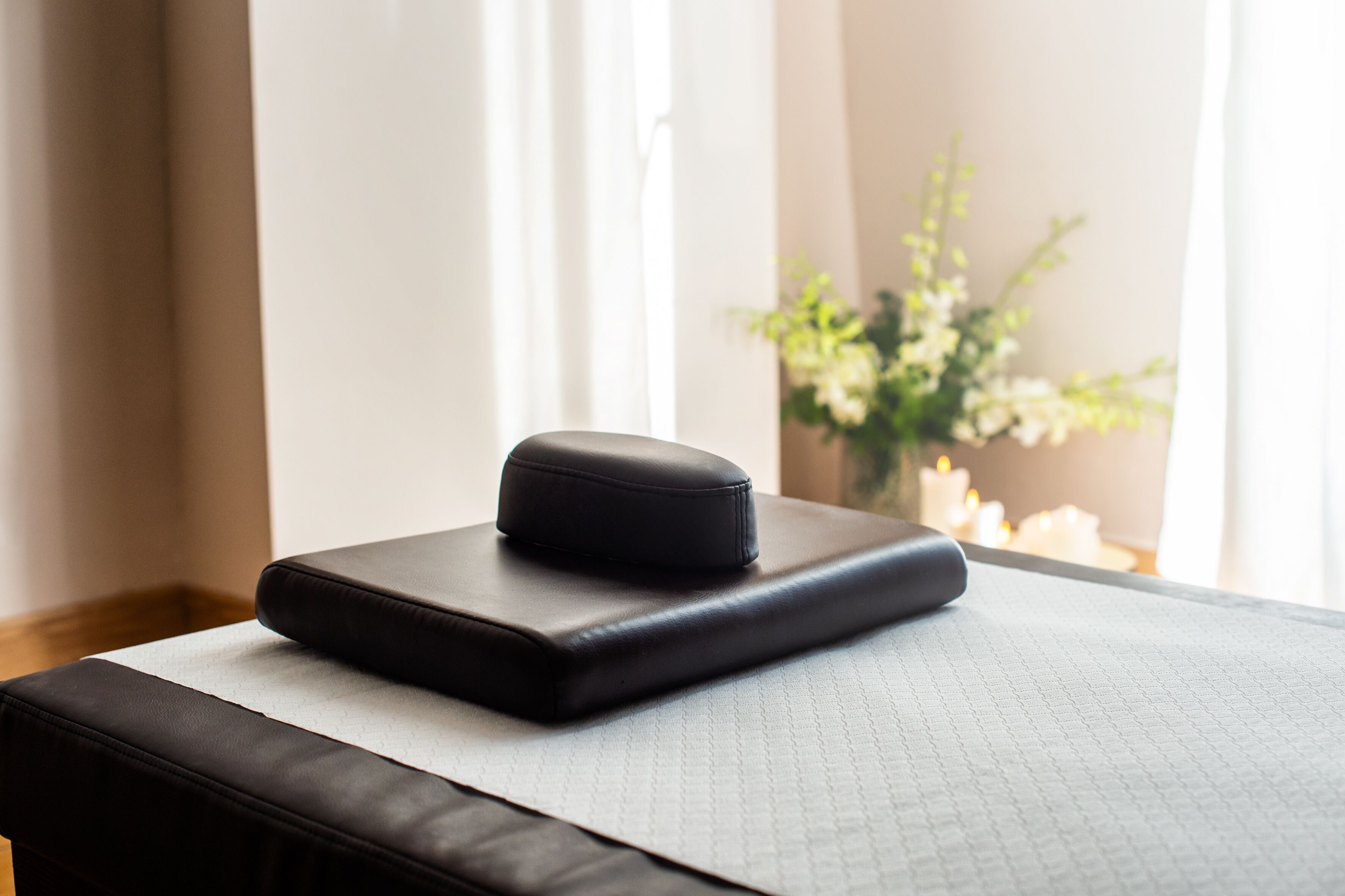 Ein Massagestuhl mit schwarzer Polsterung und strukturierter Oberfläche, platziert in der Nähe eines Fensters mit Tageslicht und einer Blumenvase.