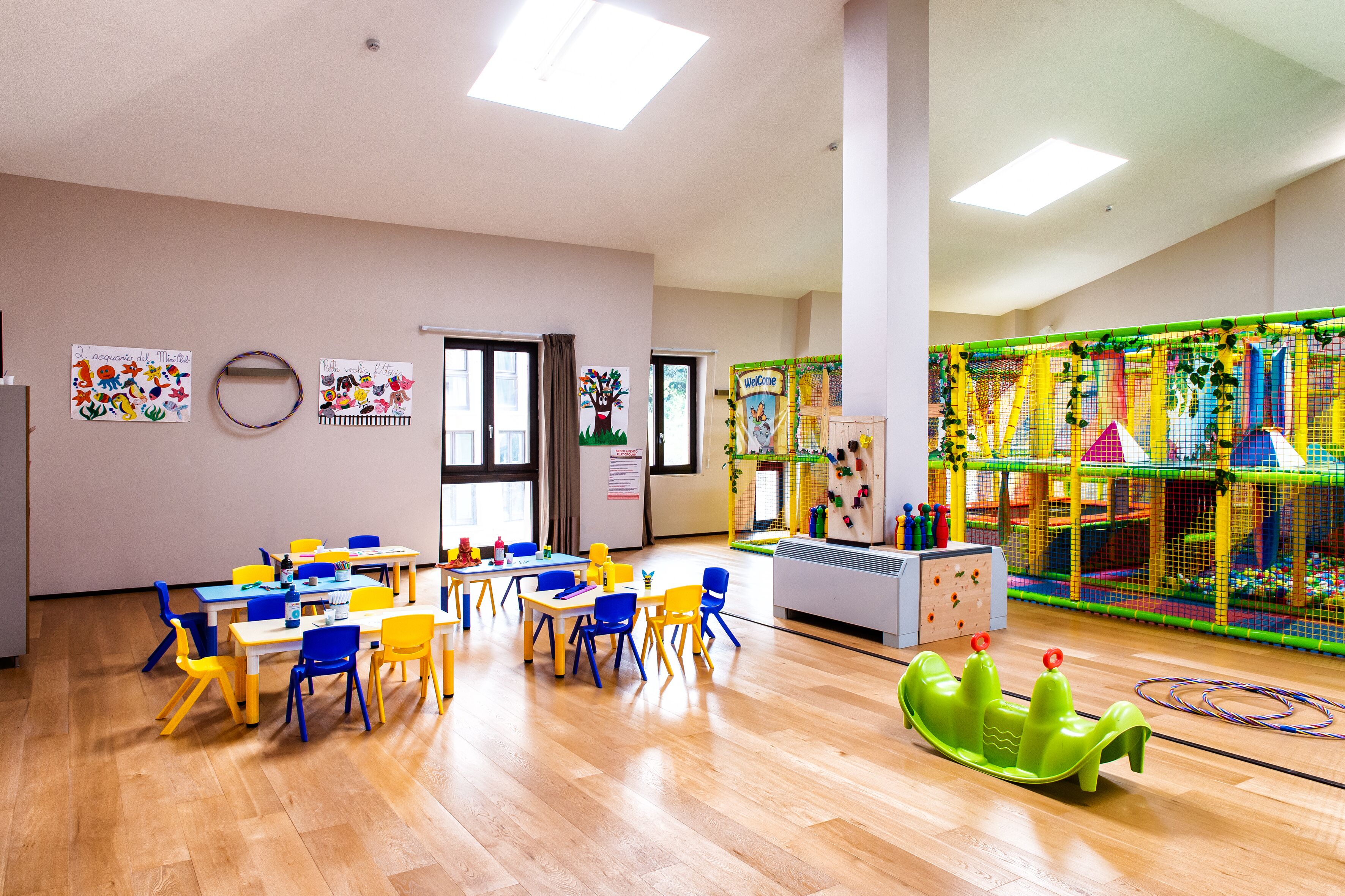 A classroom with wooden floors, yellow and blue chairs, and tables. There is a play area with colorful equipment and a green slide.