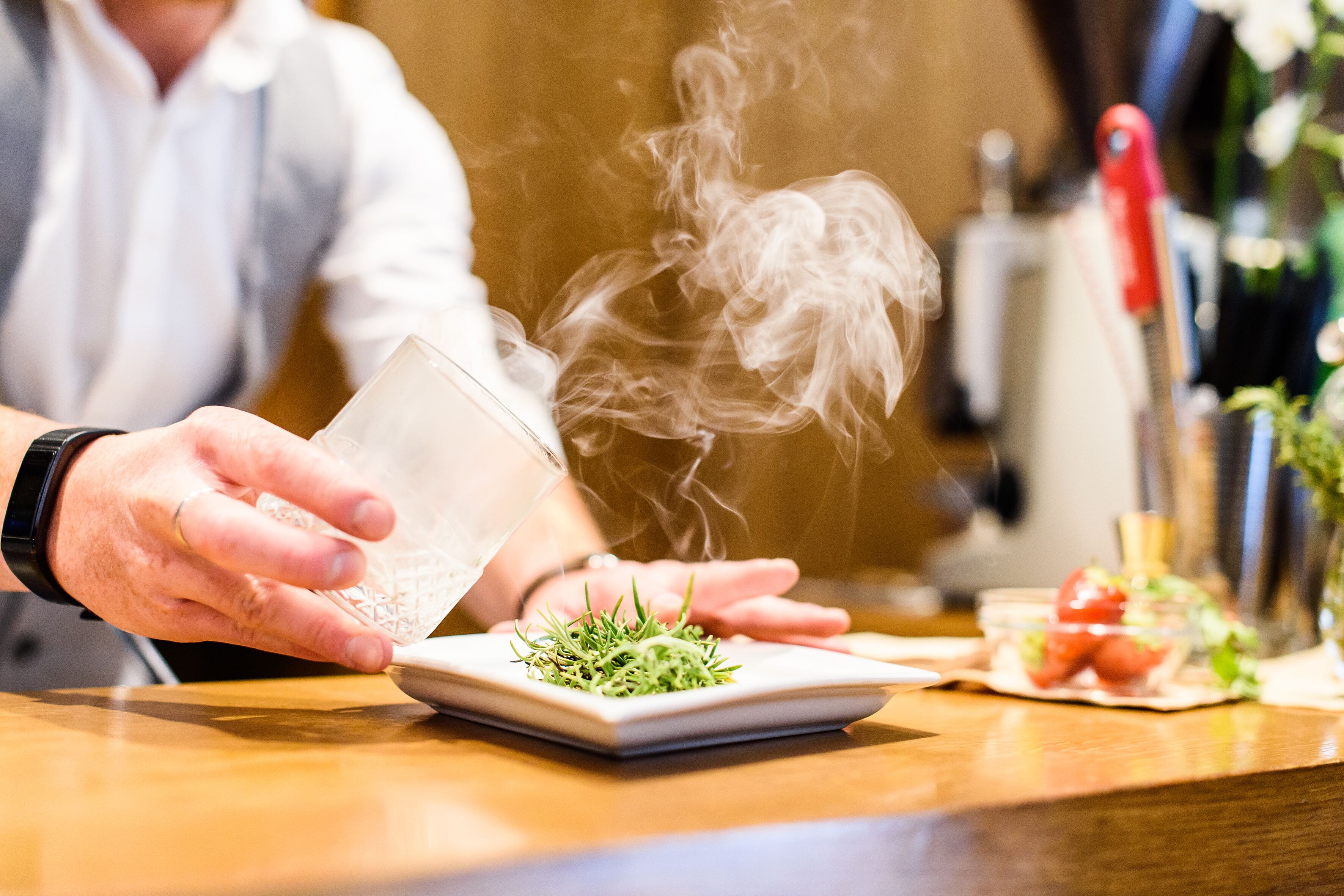 A person pouring a clear liquid onto a plate with green garnish, steam rising from the liquid.