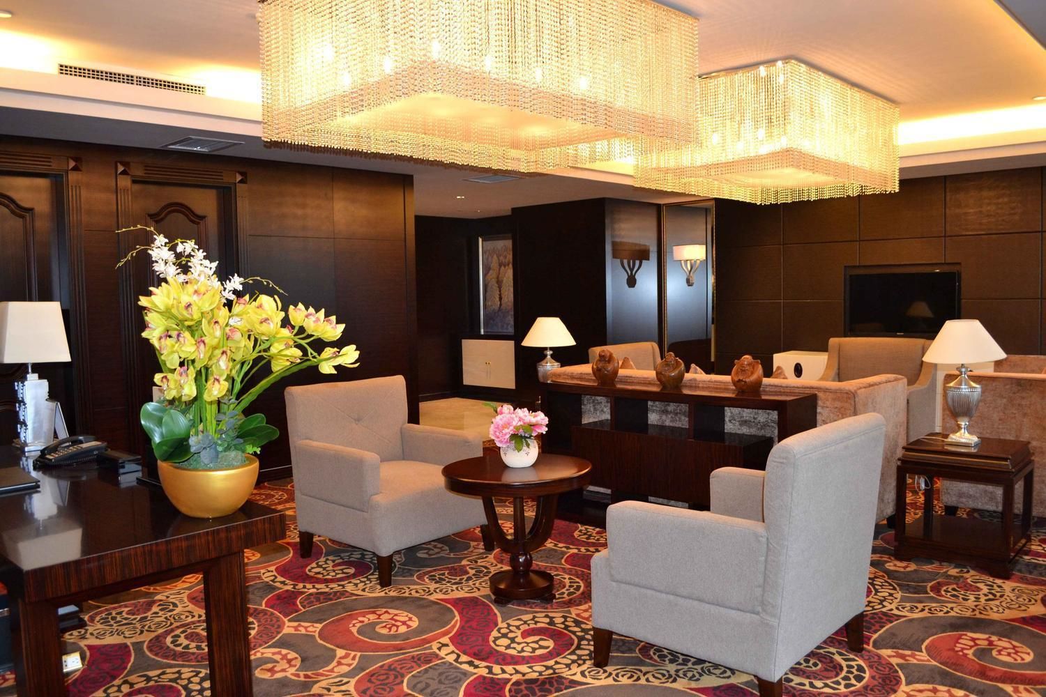 A hotel lobby with a large chandelier, comfortable seating, a reception desk, and a decorative vase with flowers.