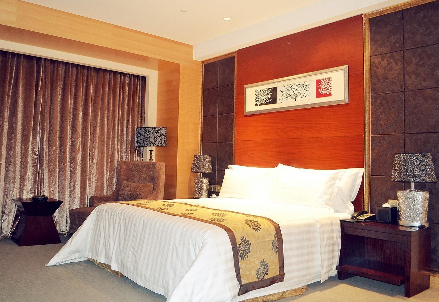 A hotel room with a large bed, two bedside tables with lamps, a chair, a large window with curtains, and a framed artwork on the wall.