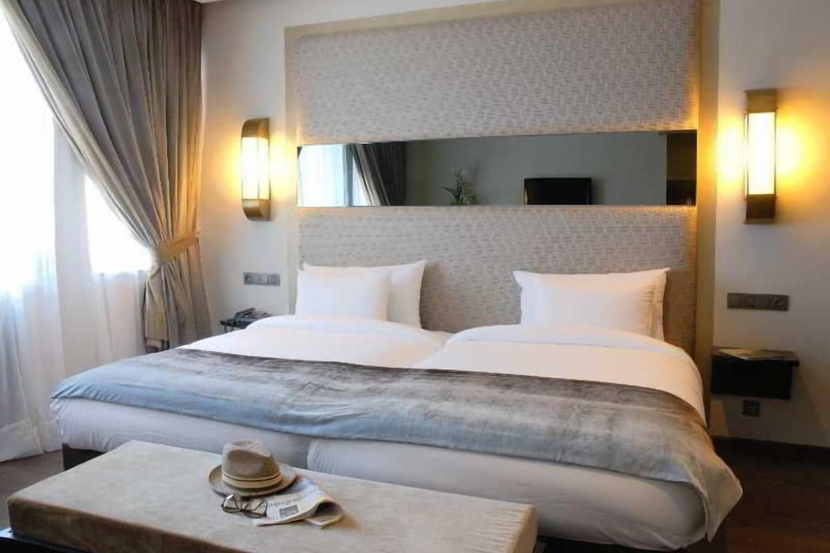 Une chambre d’hôtel avec un grand lit, deux tables de chevet, une commode, un miroir et une fenêtre avec des rideaux.