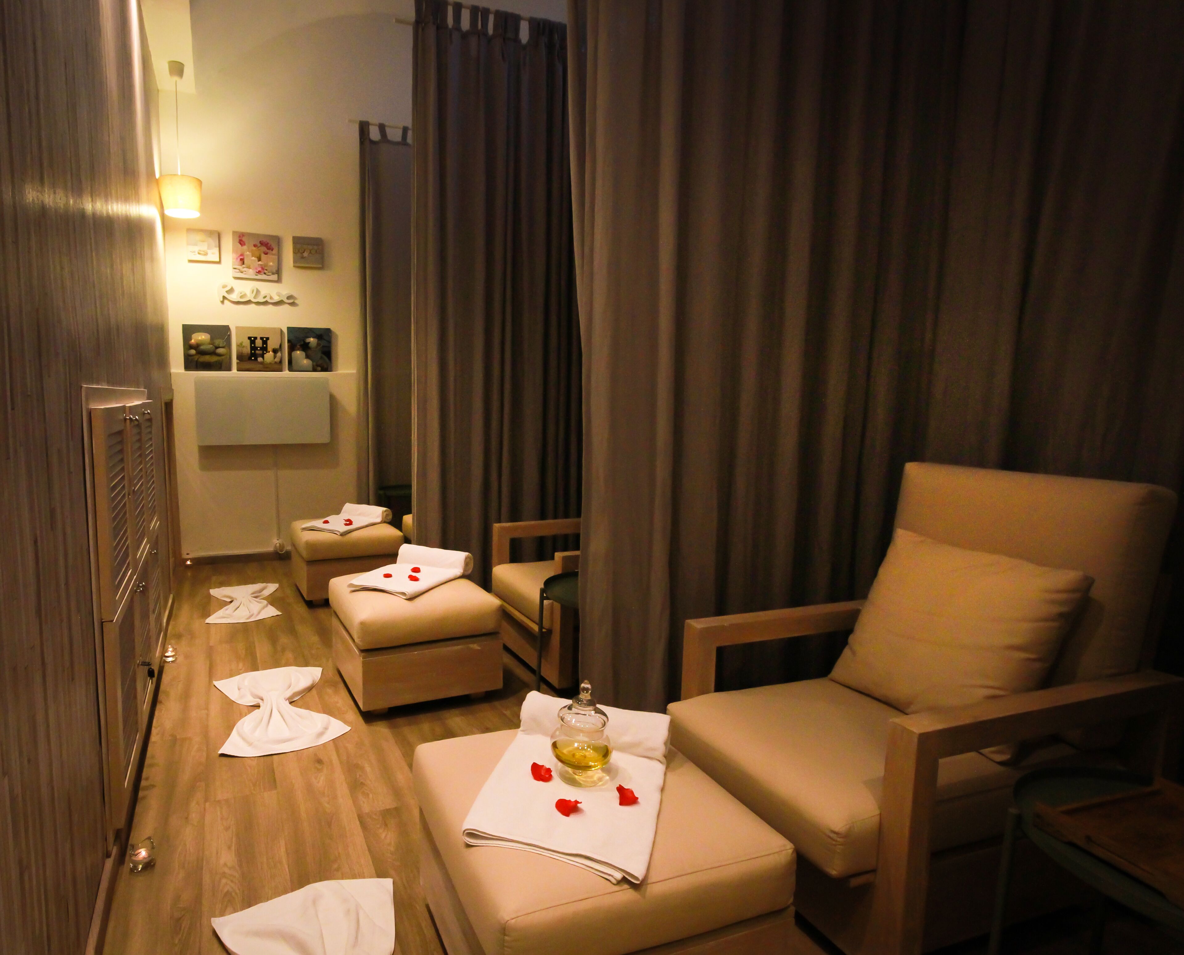 A hotel room with a wooden floor, a sofa, a chair, and a small table with a bottle and rose petals.