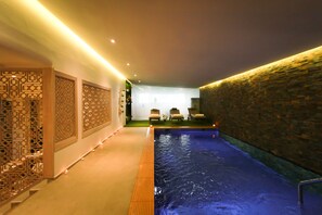 Una piscina interior con una pared de piedra en un lado y un separador decorativo en el otro. Hay sillones de descanso al fondo.