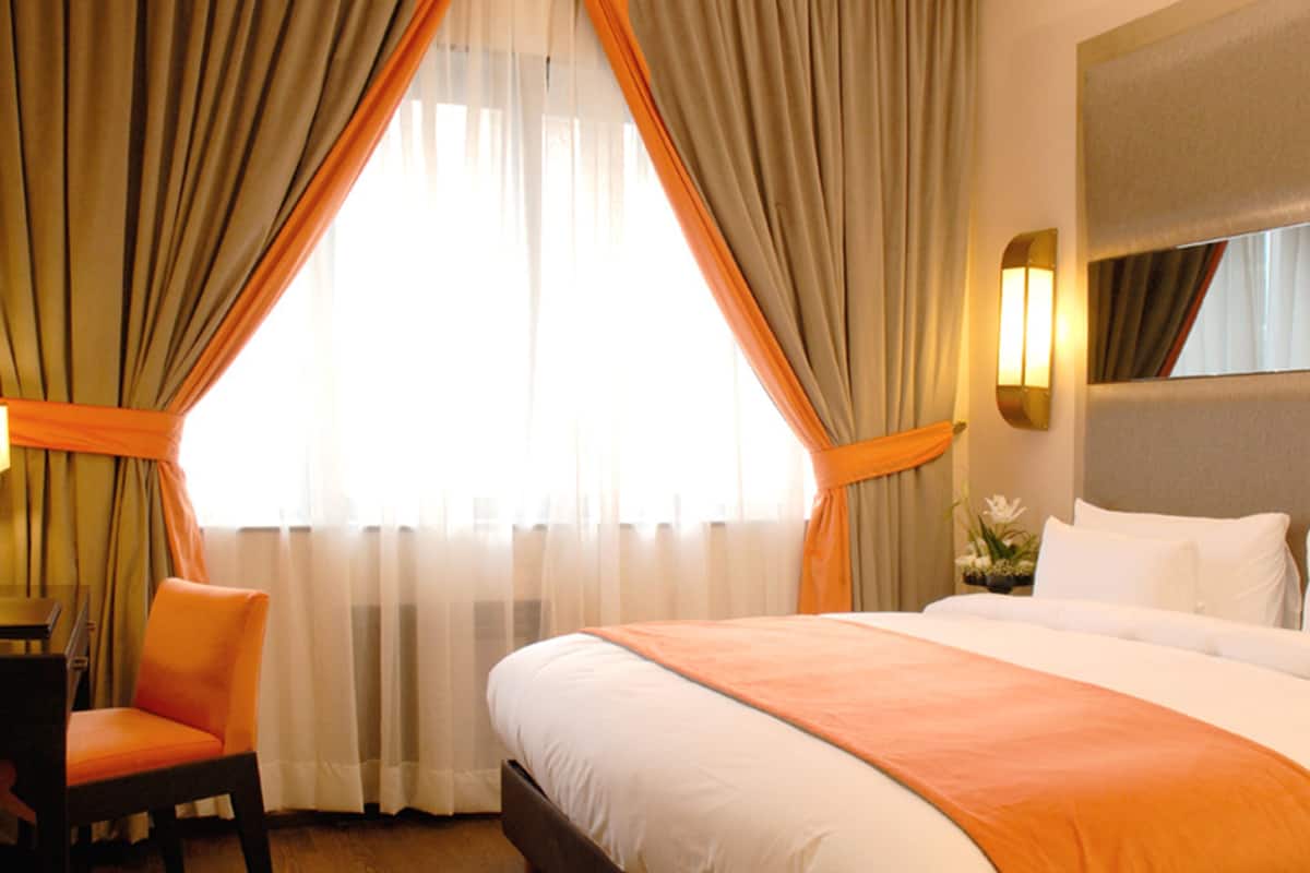 Una habitación de hotel con una cama grande, una silla anaranjada, un escritorio y una ventana con cortinas.