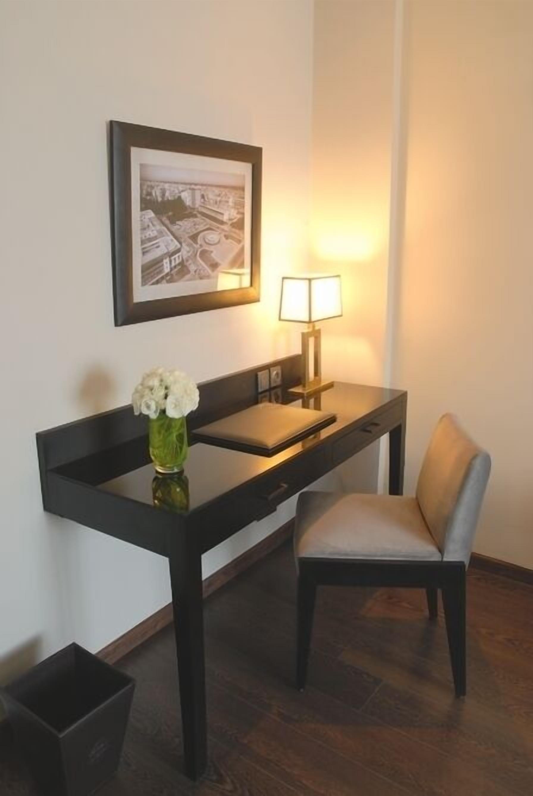 A hotel room with a desk, chair, lamp, framed picture, and a vase with flowers.