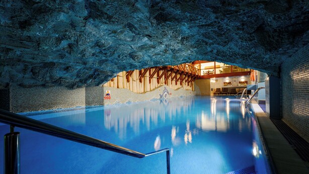 An indoor swimming pool with a rocky cave-like ceiling, a wooden deck area, and a tiled wall with a mountain scene. - Hotel Belvedere Resort & SPA (Zakopane)