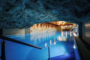 An indoor swimming pool with a rocky cave-like ceiling, a wooden deck area, and a tiled wall with a mountain scene. - Hotel Belvedere Resort & SPA (Zakopane)