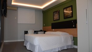 A bedroom with a bed, a desk, and a chair. There is a television mounted on the wall and a window with blinds.