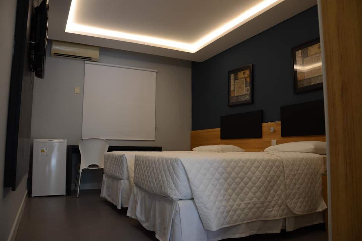 Quarto de hotel com duas camas, uma escrivaninha, uma cadeira, um quadro branco e obras de arte nas paredes.