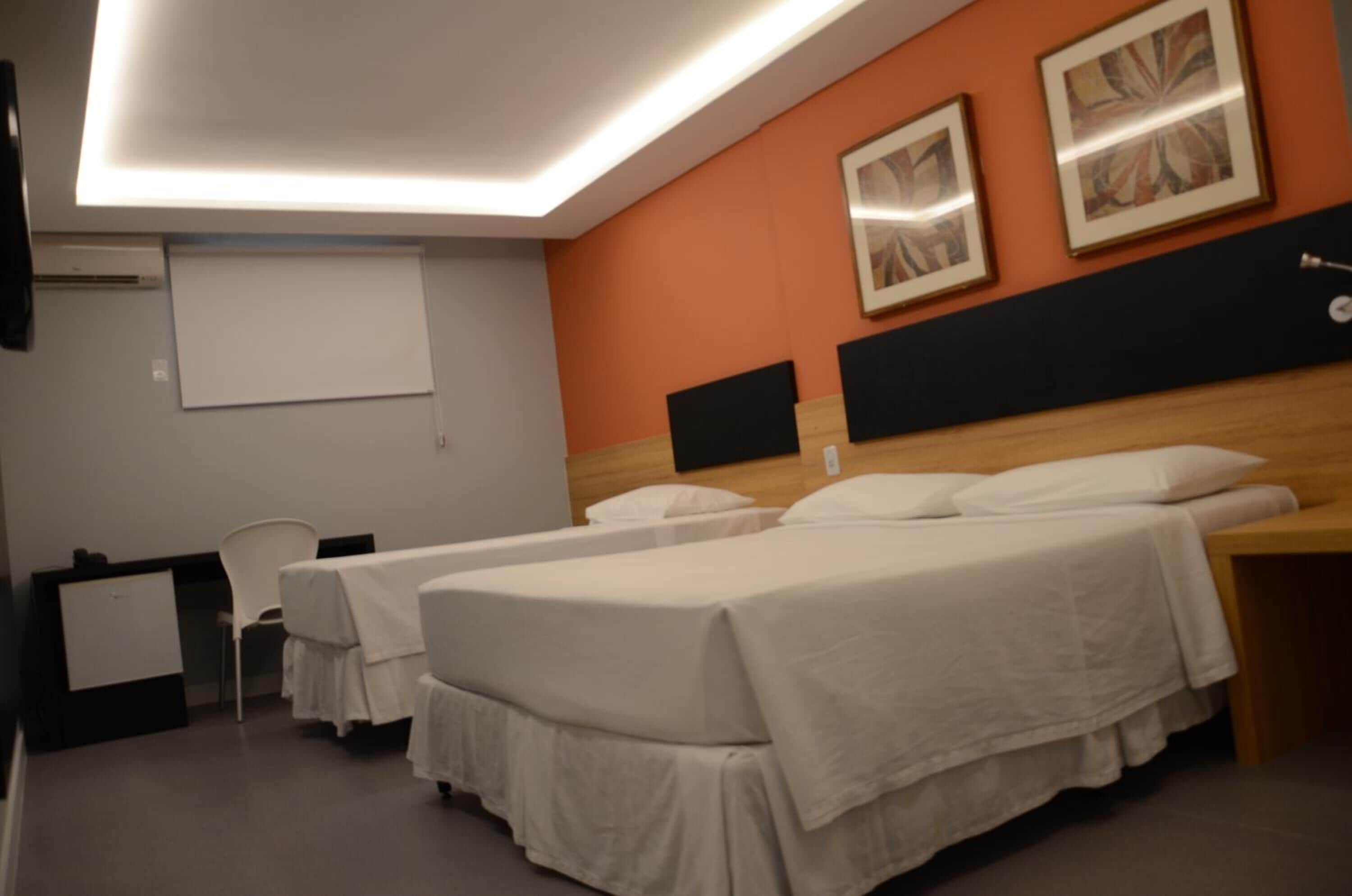 A hotel room with two single beds, a desk, a chair, and a projector screen.