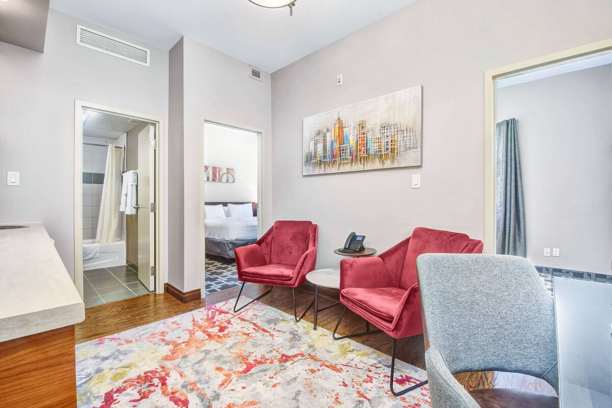 Une chambre d’hôtel moderne, équipée d’un lit, d’un bureau, de deux chaises, d’un tableau et d’une salle de bain visible à travers une porte ouverte.