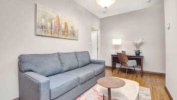 Una sala de estar moderna con un sofá gris, una mesa de centro redonda de madera, un escritorio con una silla y una pintura de un paisaje urbano en la pared.