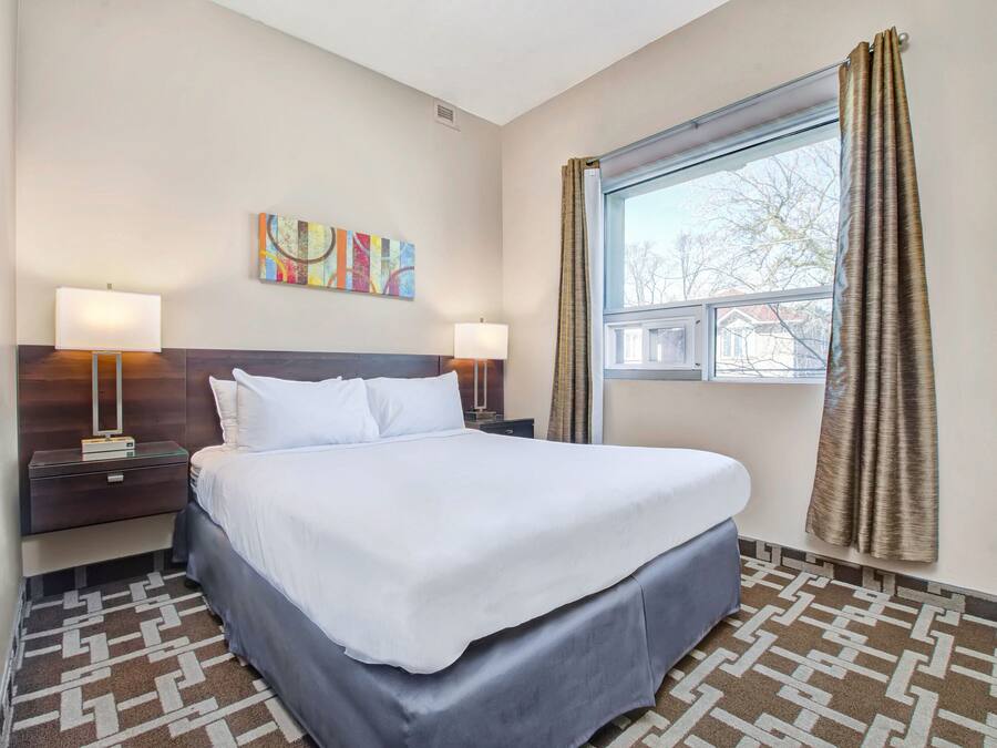 A hotel room with a large bed, two bedside tables with lamps, a colorful abstract painting, a window with curtains, and patterned carpet.