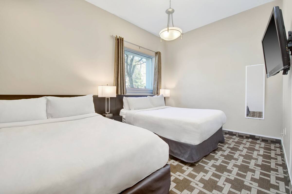 Une chambre d’hôtel avec deux lits, une grande fenêtre avec des rideaux, un téléviseur fixé au mur et un sol à motifs.