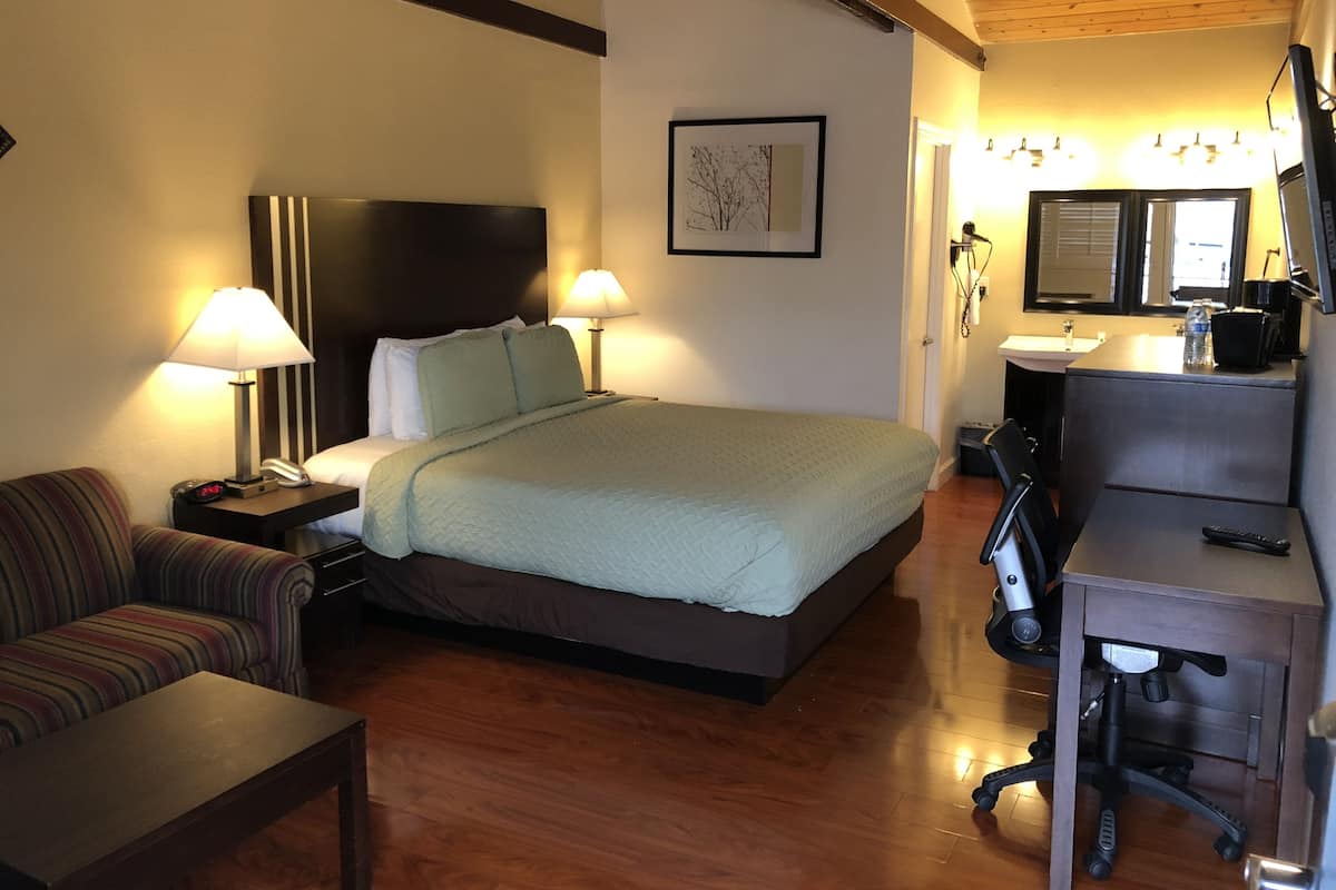 A hotel room with a bed, a desk with a chair, a sofa, a bedside table with a lamp, a wall-mounted TV, and a bathroom with a sink and mirror.
