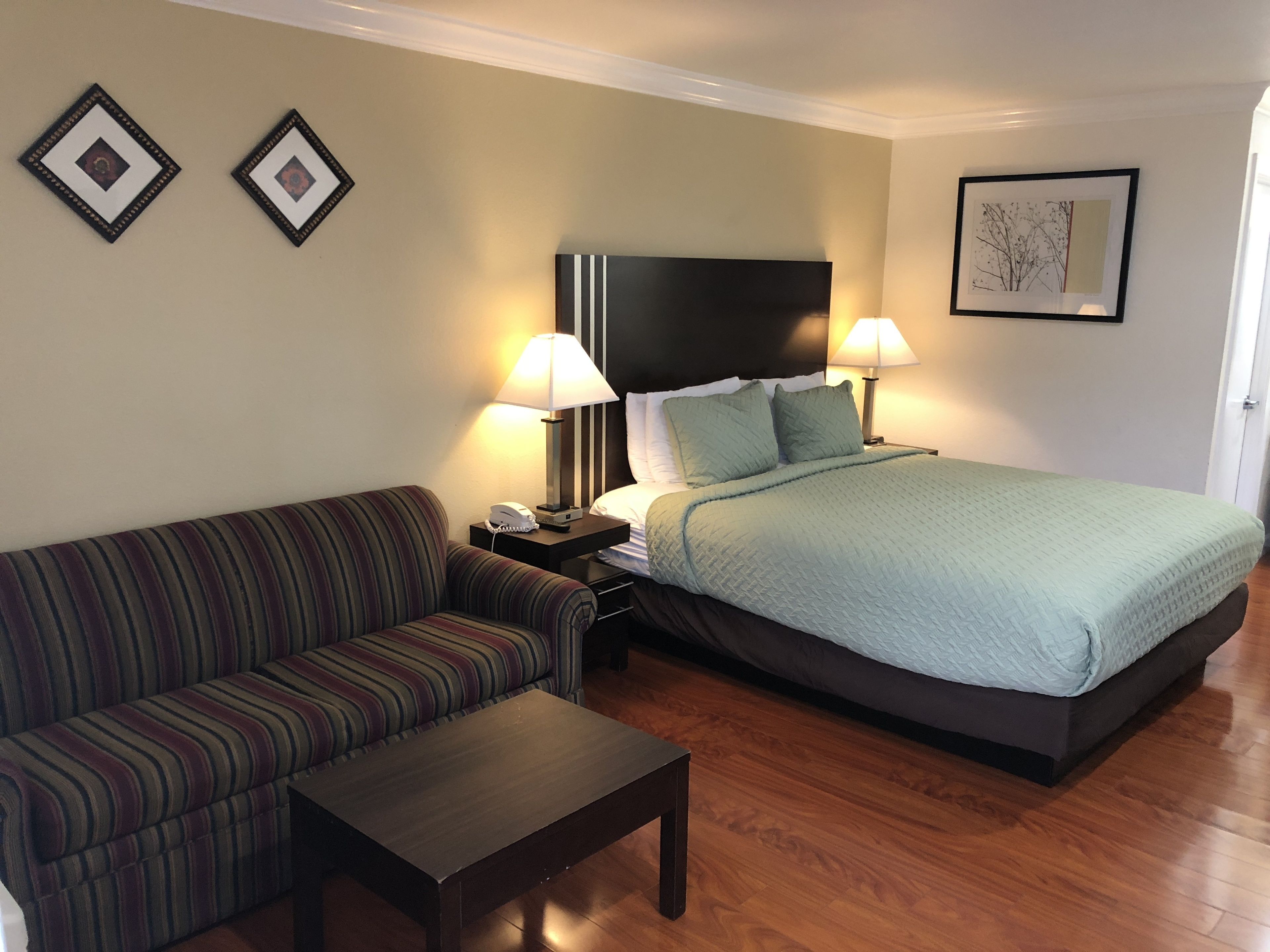 A hotel room with a striped sofa, a single bed with a green bedspread, two bedside lamps, a nightstand with a phone, and two framed pictures on the wall.