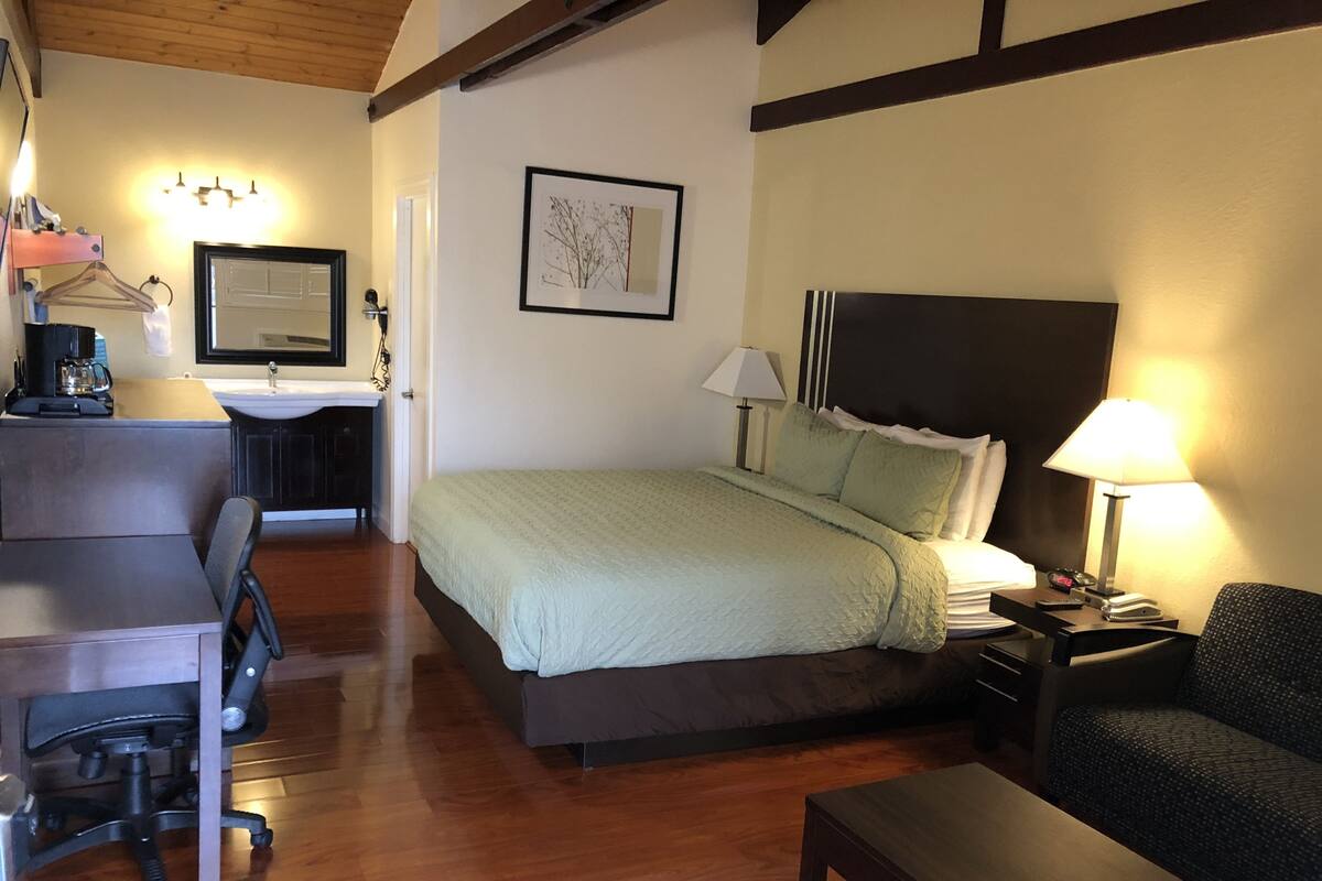 A hotel room with a bed, bedside table, lamp, chair, desk, and a small kitchenette area.