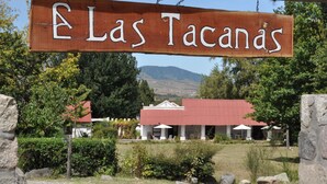 Ein Holzschild mit der Aufschrift 'Las Tacanas', mit Blick auf ein Gebäude mit rotem Dach und weißen Säulen, umgeben von Grün und Hügeln im Hintergrund.