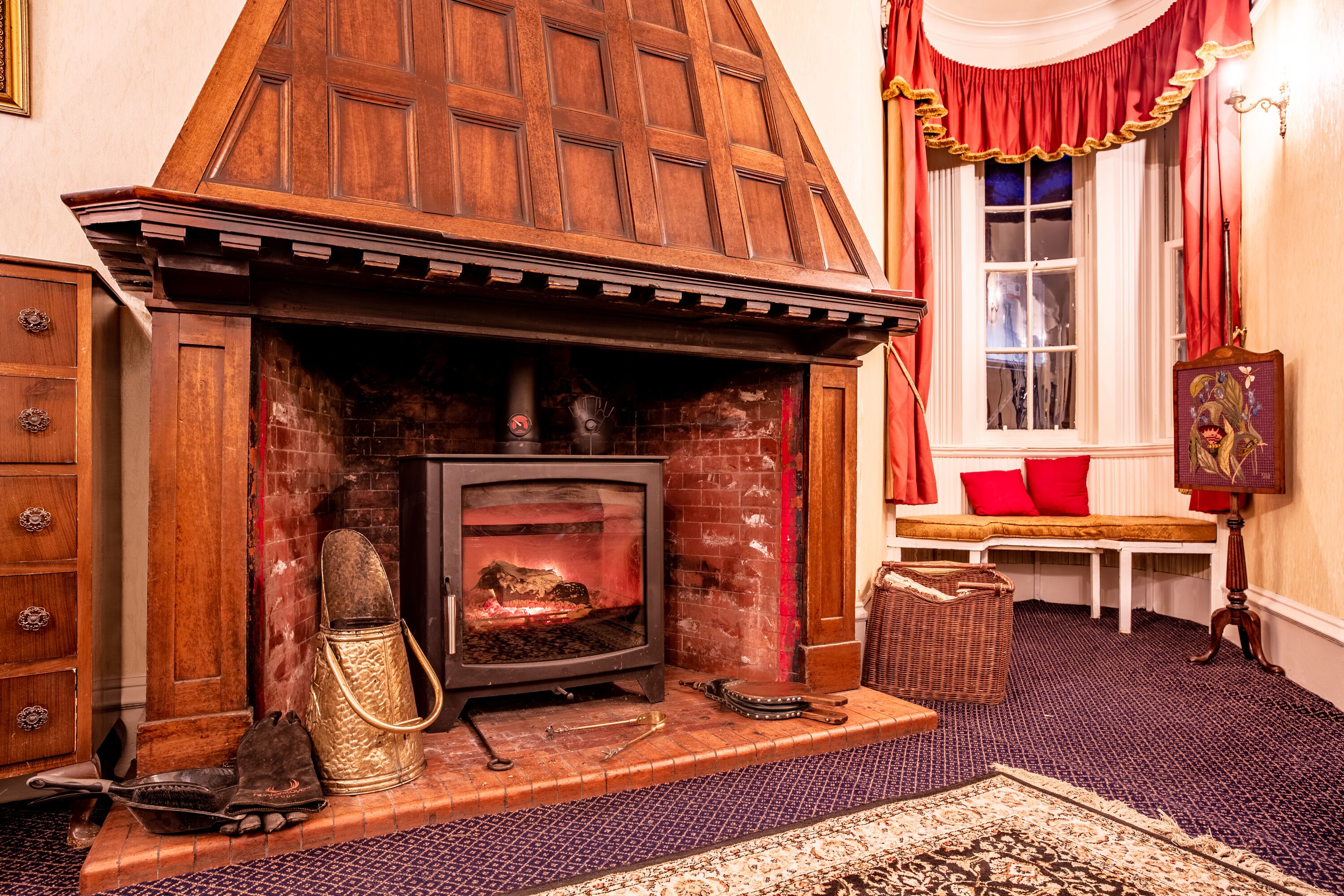 A traditional fireplace with a wooden mantel, a bench with red cushions, and a wicker basket.