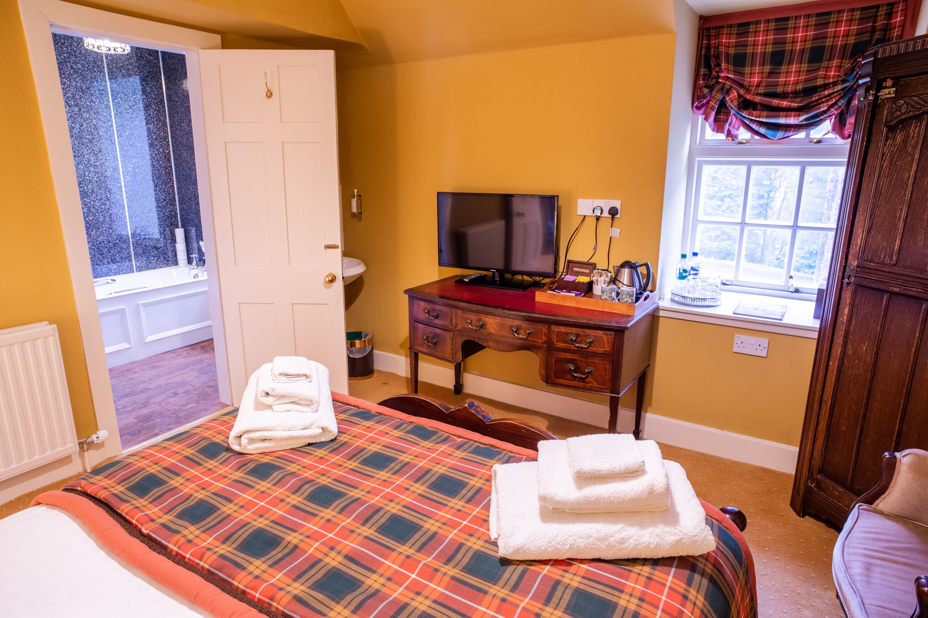 Habitación de hotel con edredón a cuadros, televisión, cómoda de madera y ventana con cortina a cuadros.