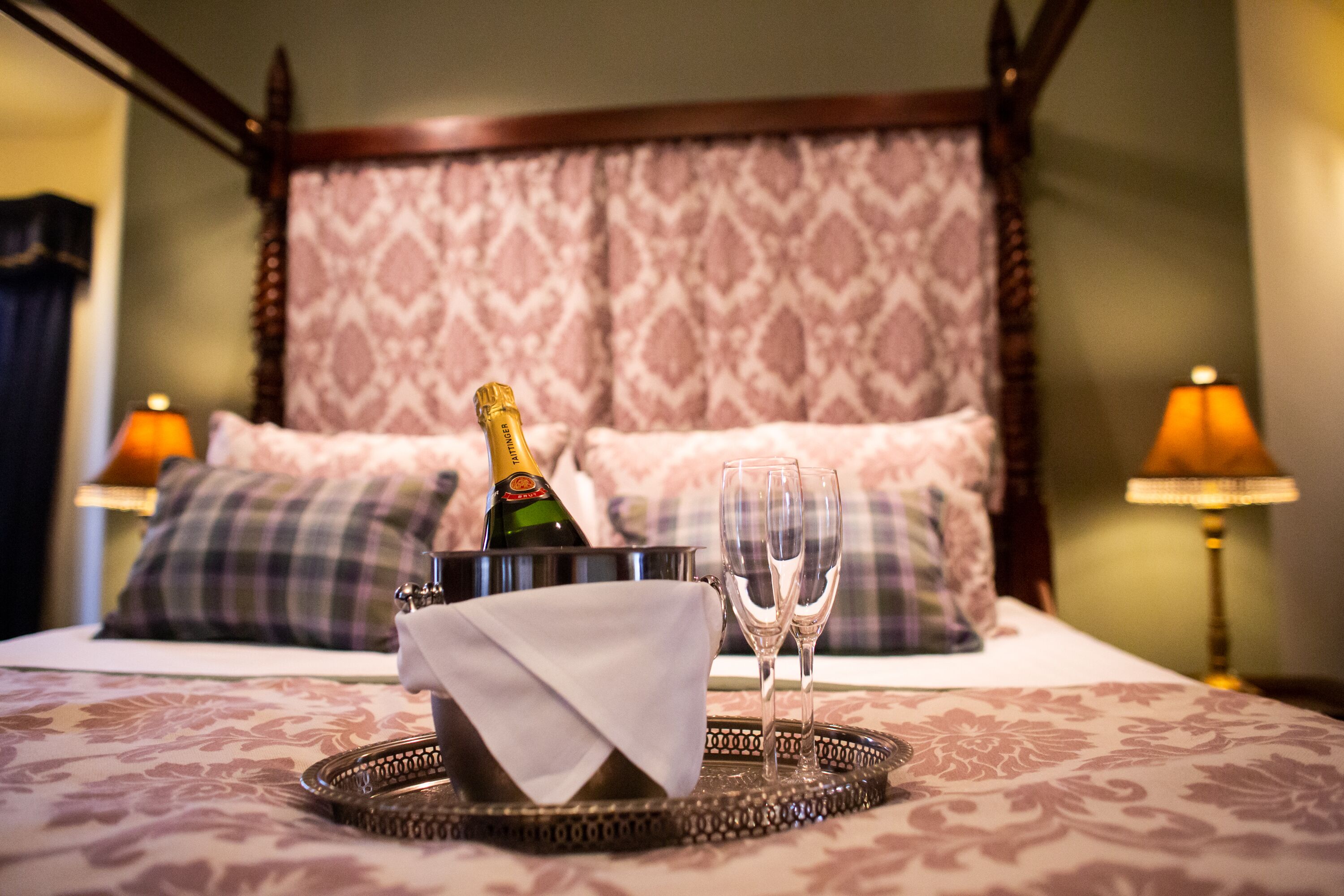 A bed with a decorative headboard, a tray with a bottle of champagne and two glasses, and two lamps on either side.