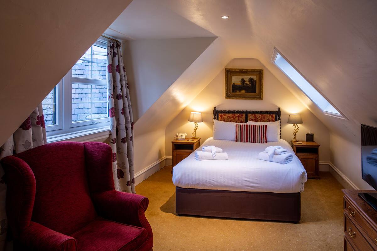 Une chambre à coucher avec un grand lit, deux tables de chevet avec des lampes, un fauteuil rouge et une fenêtre avec des rideaux.