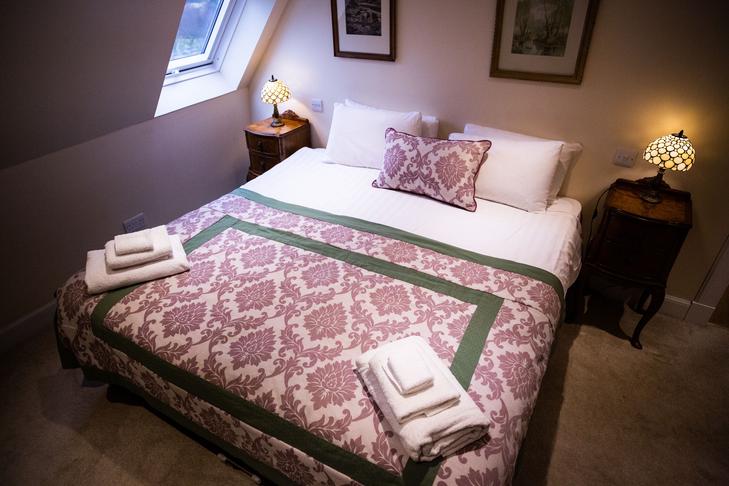 A neatly made bed with a floral bedspread, two bedside lamps, and a window with a view.