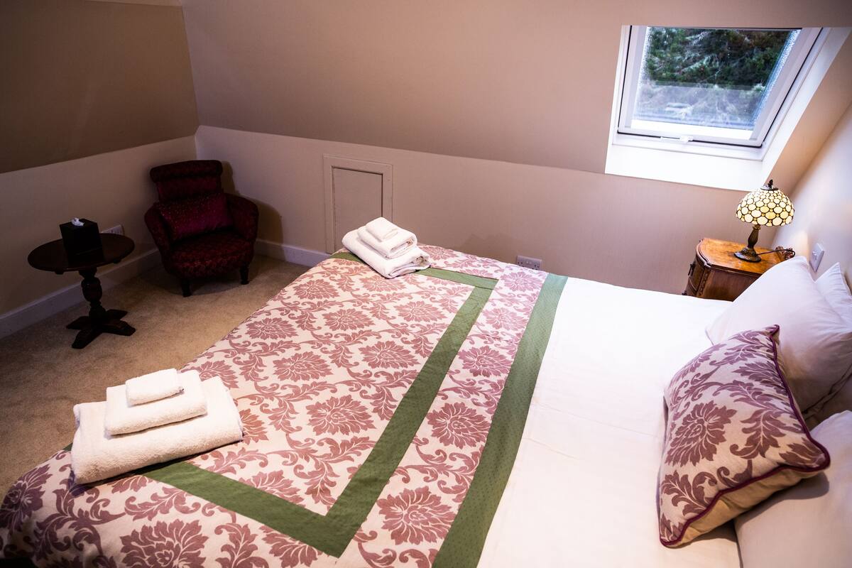 Un lit bien fait, recouvert d’une couvre-lit à motifs floraux, agrémenté d’un chemin de table vert et de serviettes pliées. On y trouve également un fauteuil rouge, une petite table avec une lampe et une fenêtre donnant sur un paysage verdoyant.
