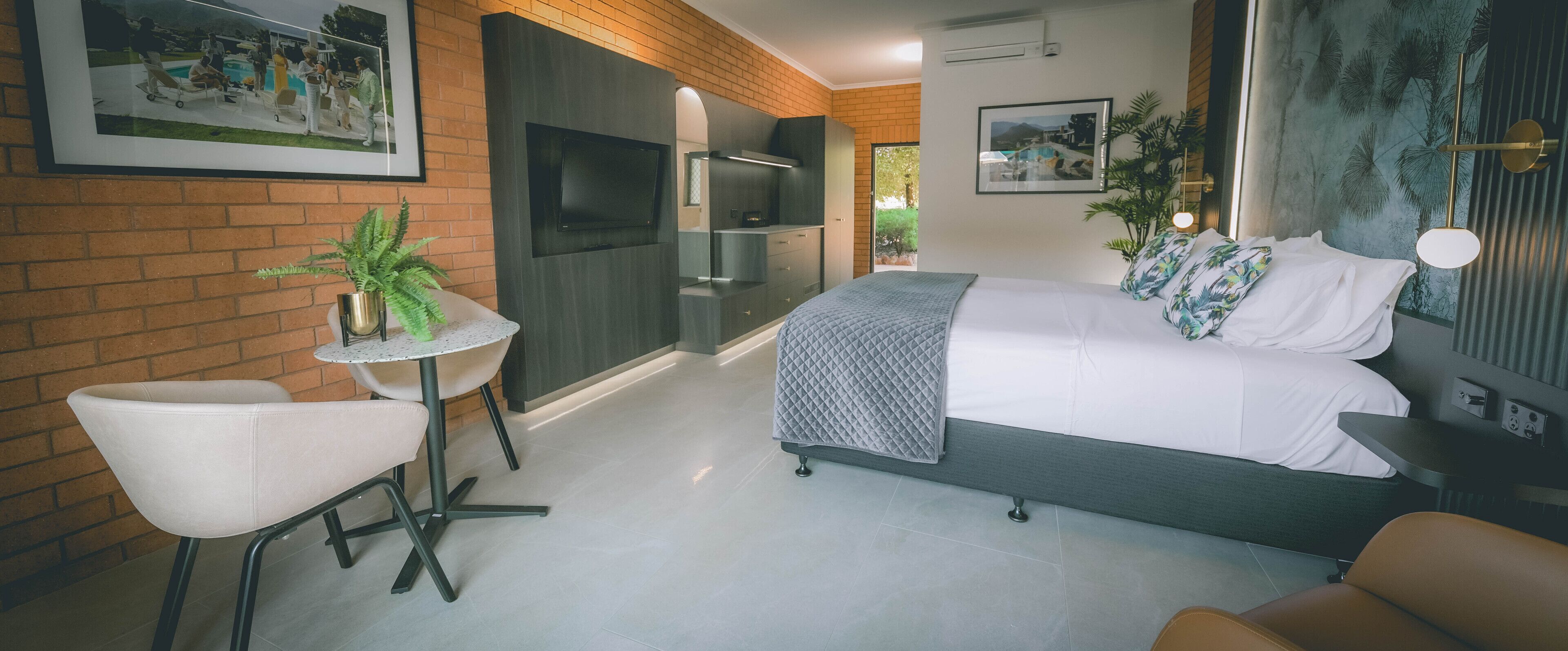 A modern hotel room with a bed, a small table, a chair, a TV, and a plant.