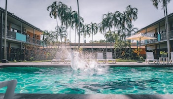 Una piscina con fuente de agua, rodeada de sillas de descanso y palmeras, con edificios al fondo.