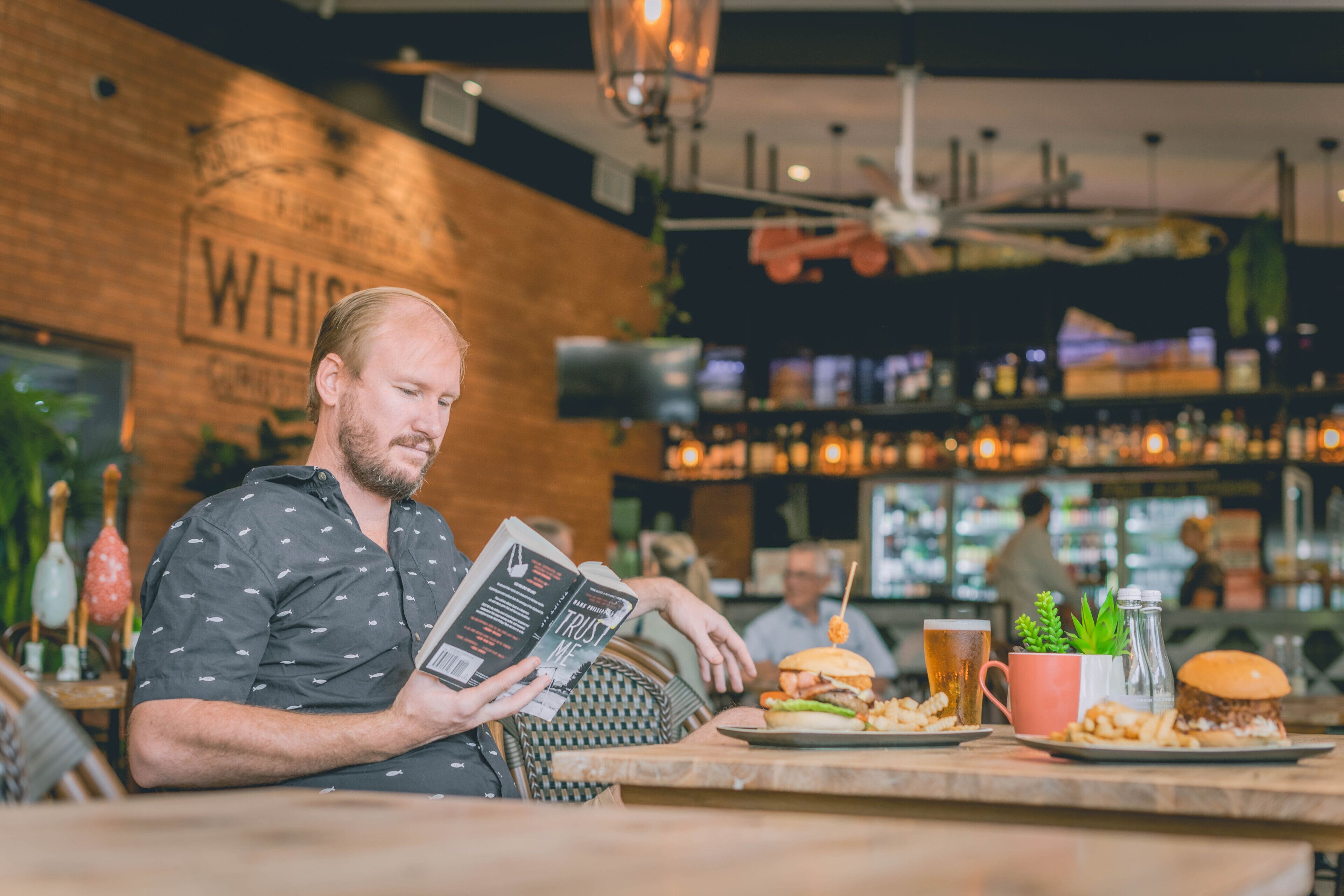 A man reading a book at a table with burgers and a beer.