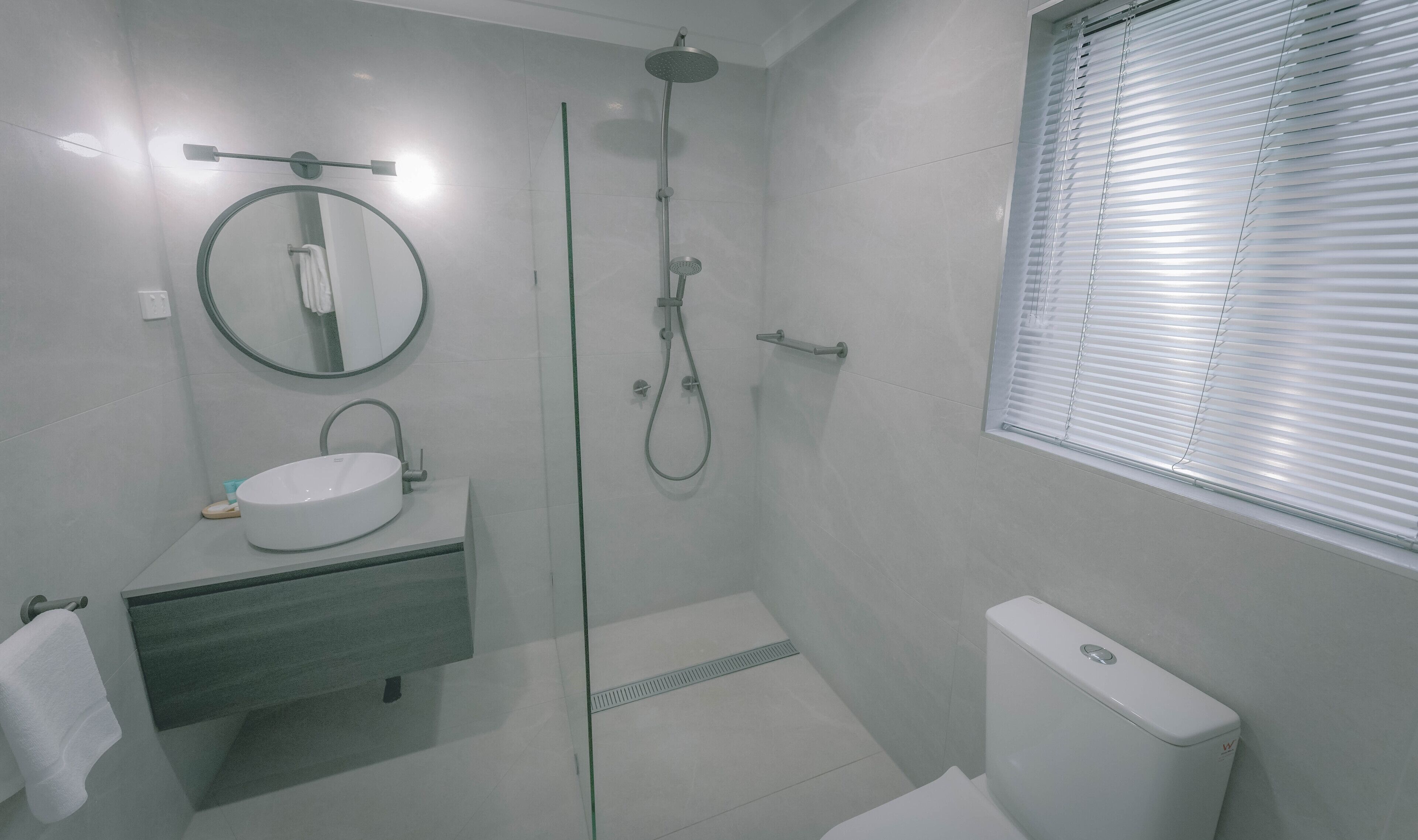 Casa de banho moderna com chuveiro envidraçado, espelho redondo, lavatório branco e sanita.