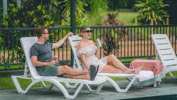 Two people relaxing on sun loungers by a poolside, with a table and a watermelon-themed tablecloth nearby.
